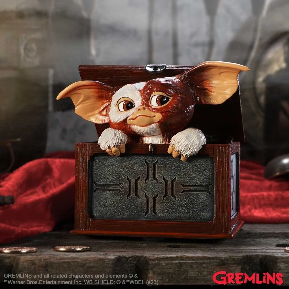 A Gremlins figurine of Gizmo, a small creature with large ears and a cute face, resting on an open wooden box with a metal emblem. The background features a rustic setting with a red cloth and coins on a wooden surface.