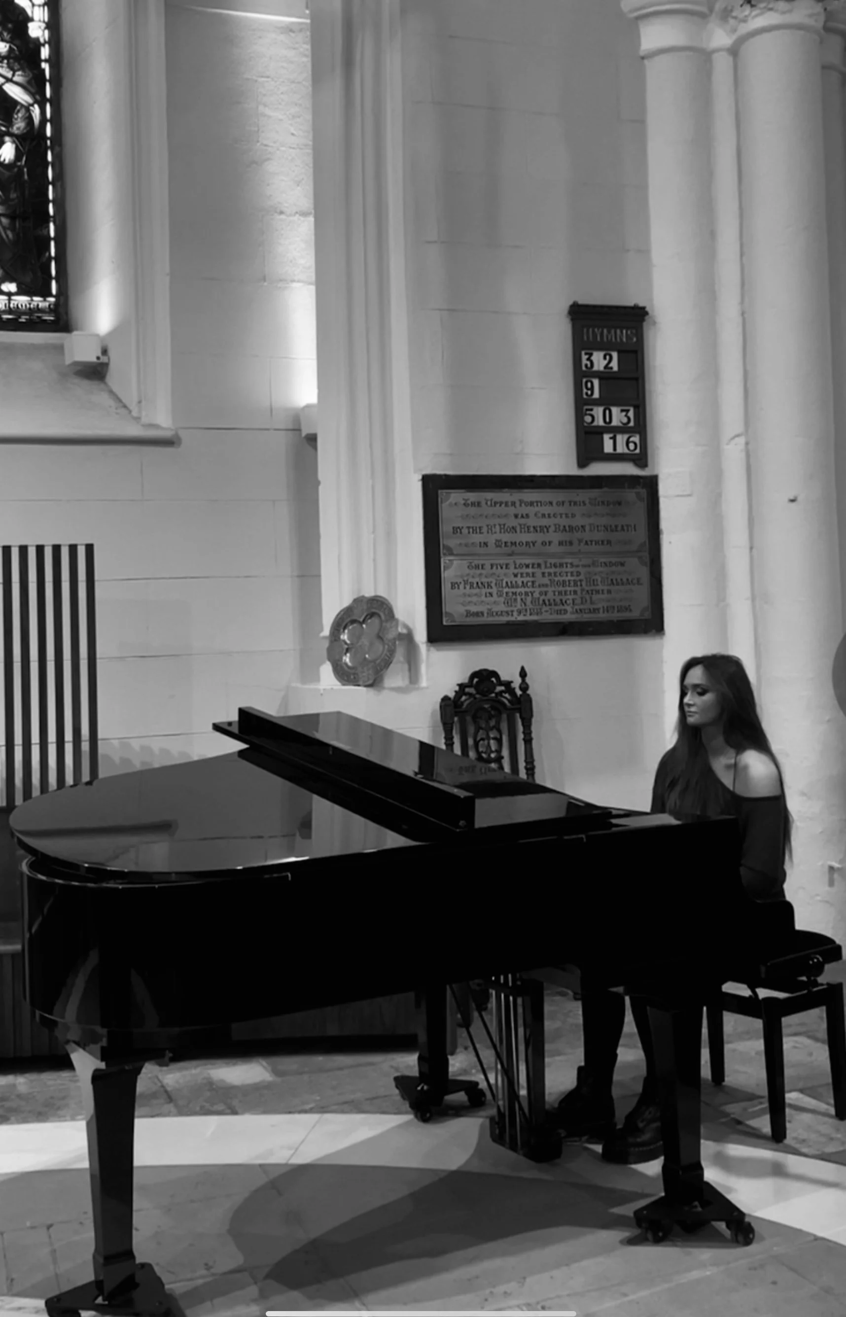 A young woman playing a grand piano inside a church with tall columns and stained glass windows, in black and white.
