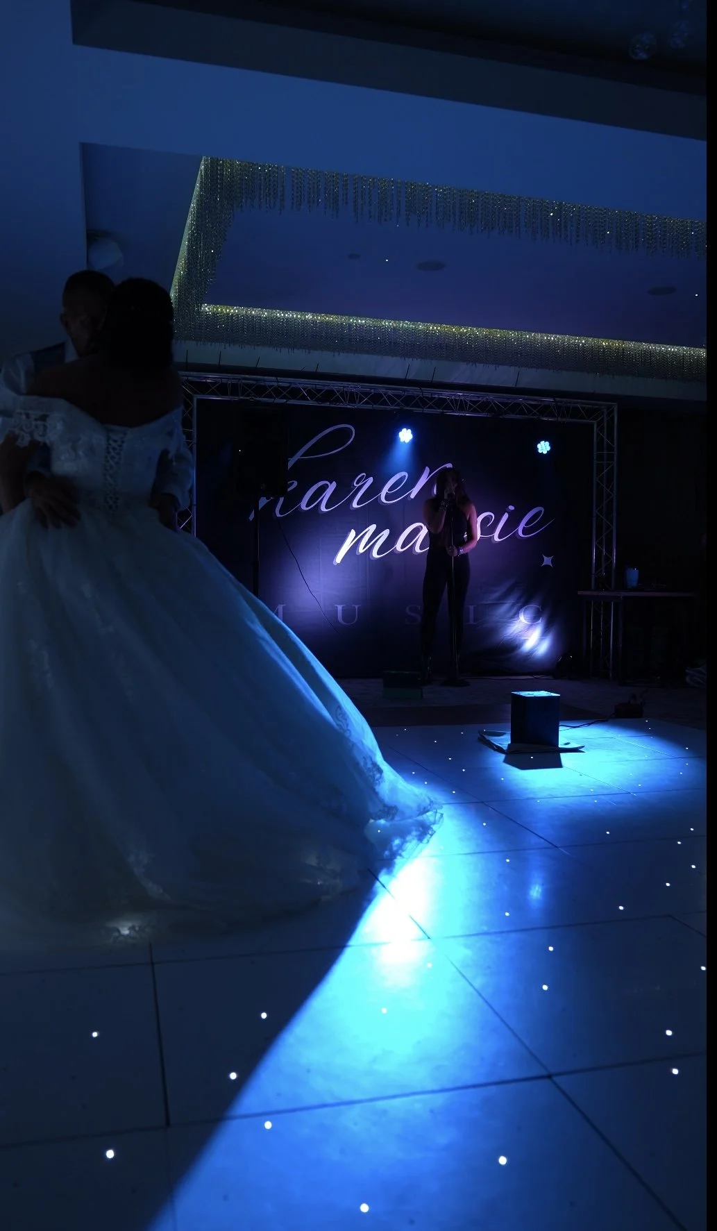 Nighttime scene at a wedding reception with a bride in a white gown dancing, a woman singing on stage in the background, and a decorated backdrop with the name 'Charen Marie' illuminated by stage lights.