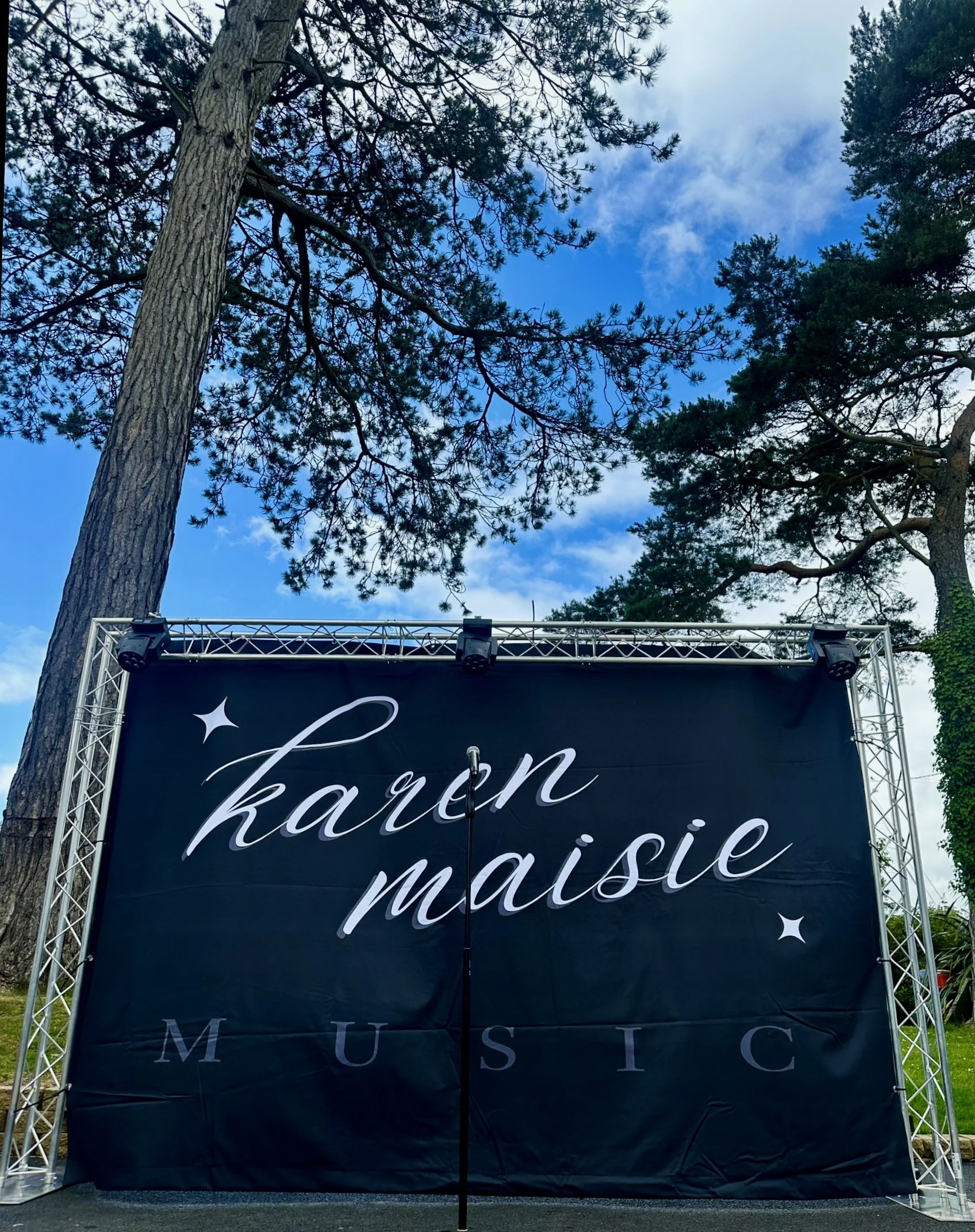 Outdoor stage with a black backdrop displaying the words 'karen maisie MUSIC' in white cursive letters, surrounded by tall trees and a partly cloudy sky