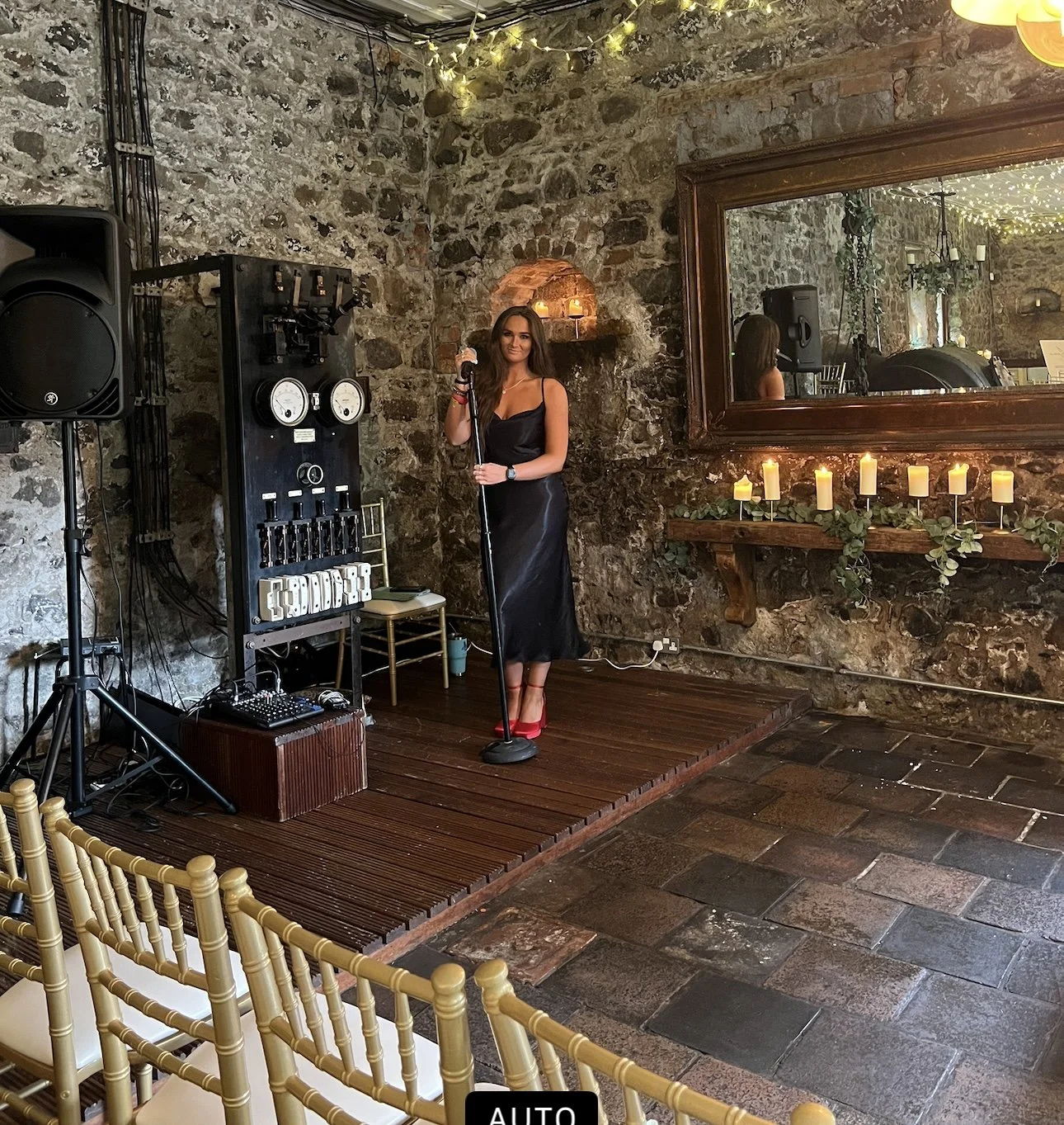 A woman in a black dress and red heels standing on a small wooden stage in a rustic venue with stone walls, holding a microphone. Behind her are musical equipment, a large mirror, and lit candles on a shelf with greenery. Empty golden chairs are in t