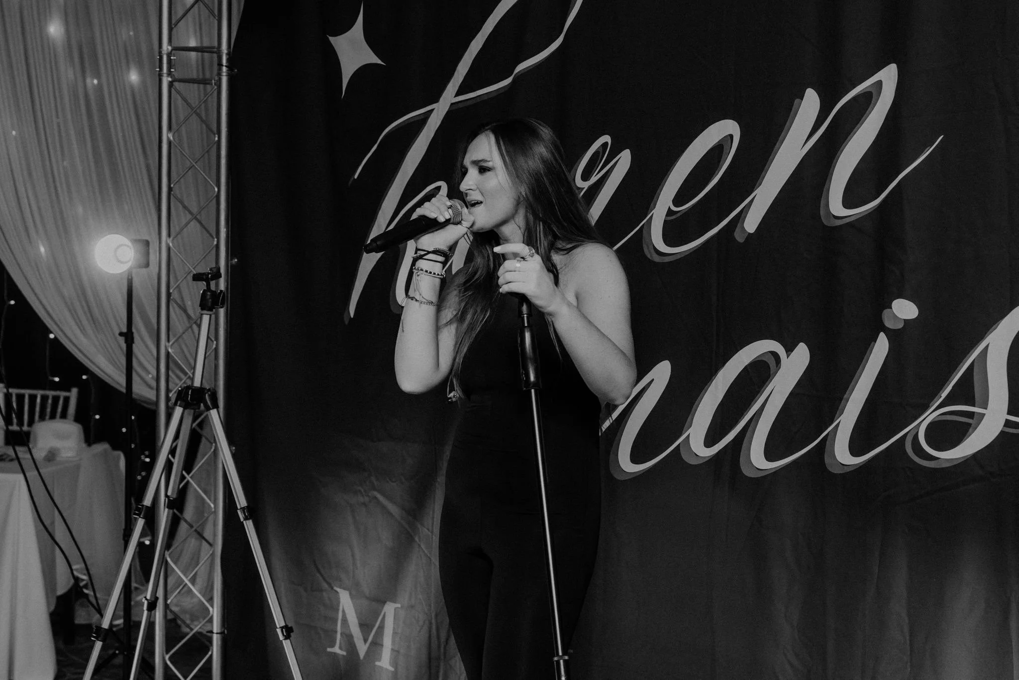 A woman singing into a microphone on stage during a talent event, with a backdrop that reads 'Talent Americas'.