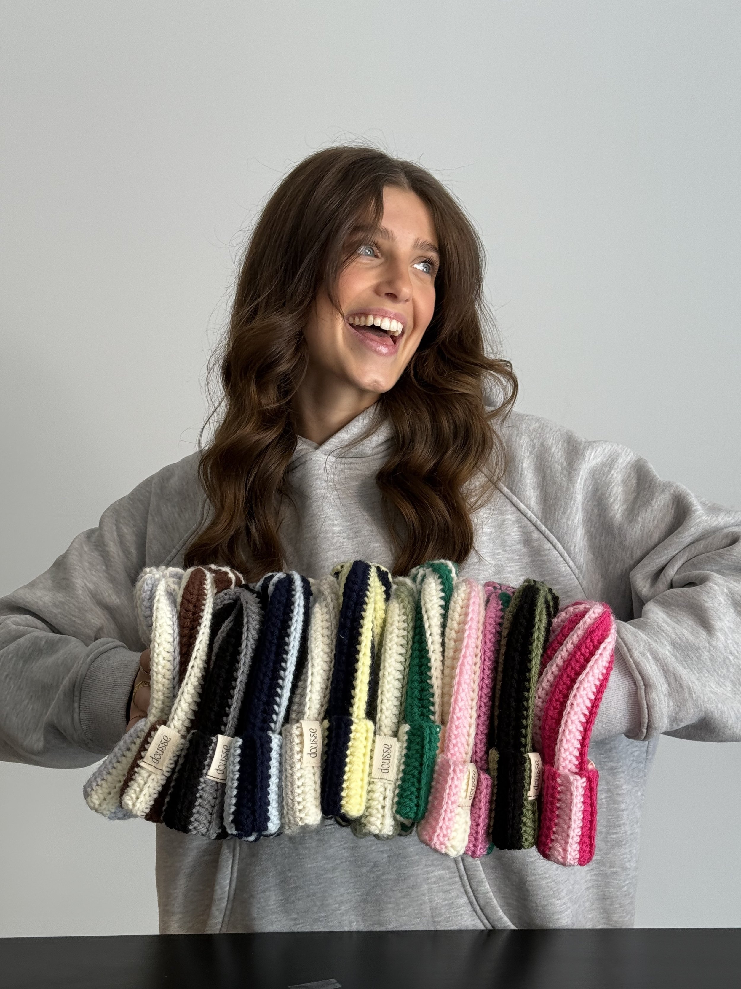 Une femme souriante tient une rangée de moufles tricotées de différentes couleurs. Elle porte un sweat gris clair et se trouve devant un fond neutre.