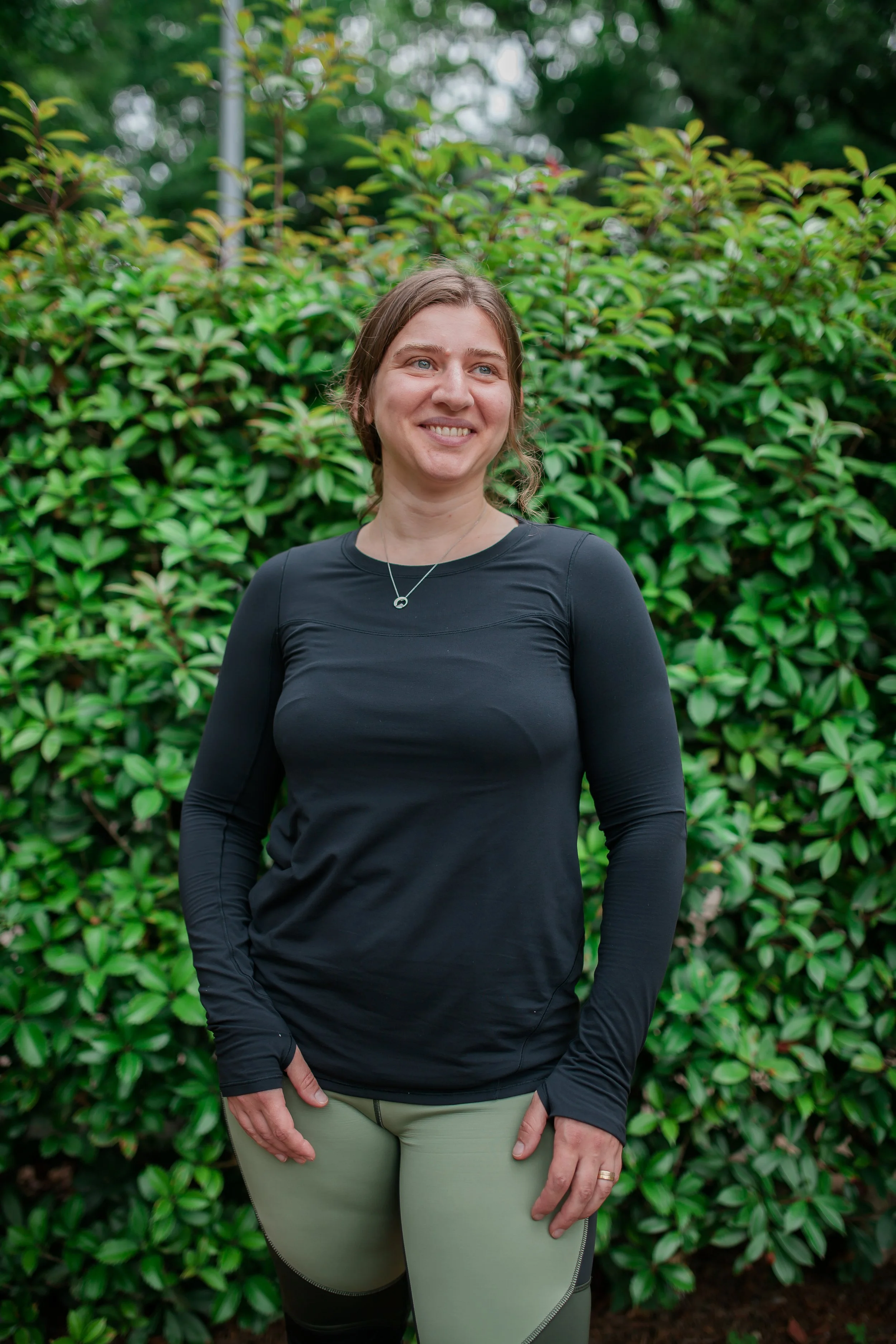 Woman standing outdoors in front of green bushes, smiling, wearing a black long-sleeve athletic shirt and beige pants.