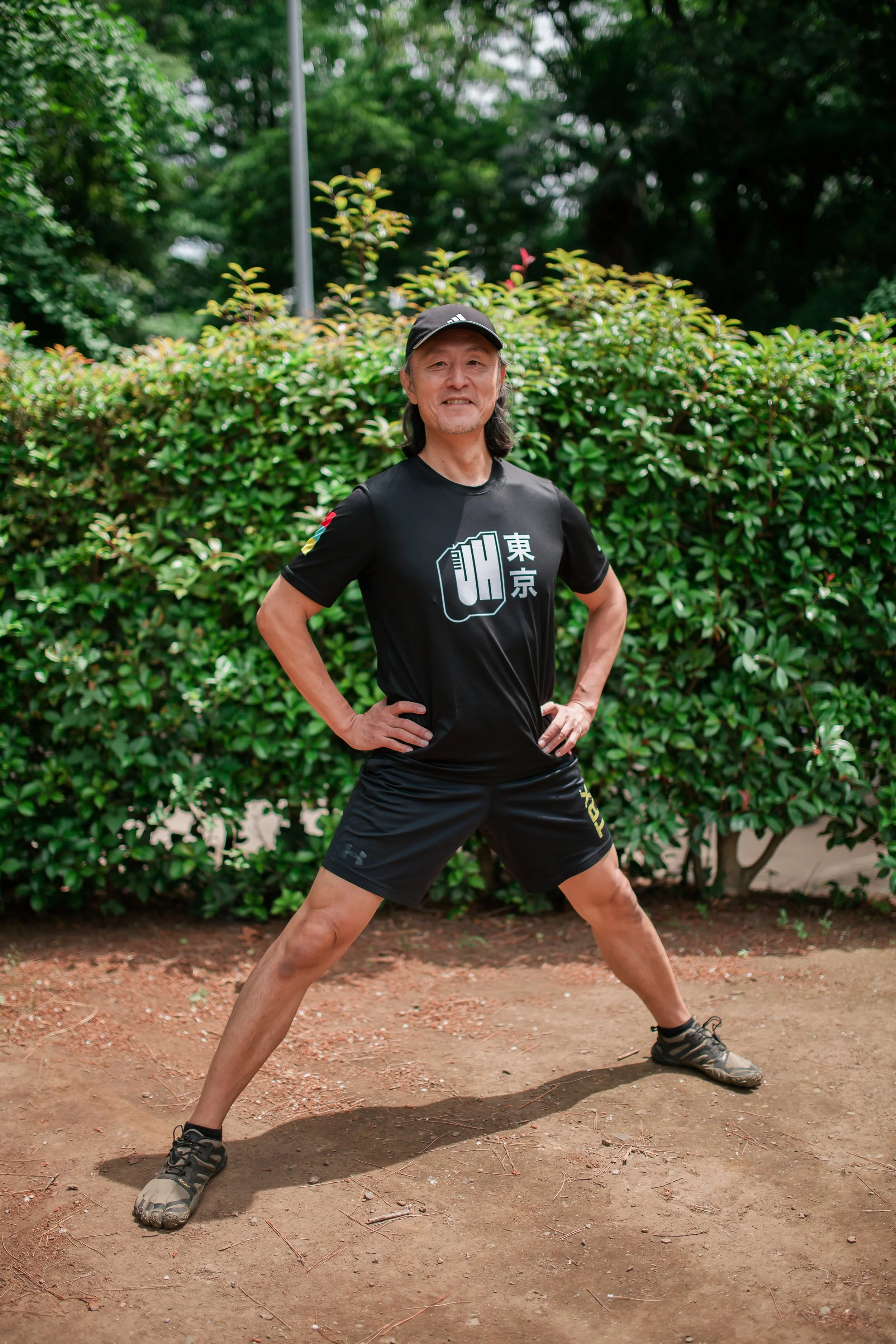 A man in athletic clothing standing with legs wide apart and hands on hips on a dirt path, with green bushes and trees in the background.