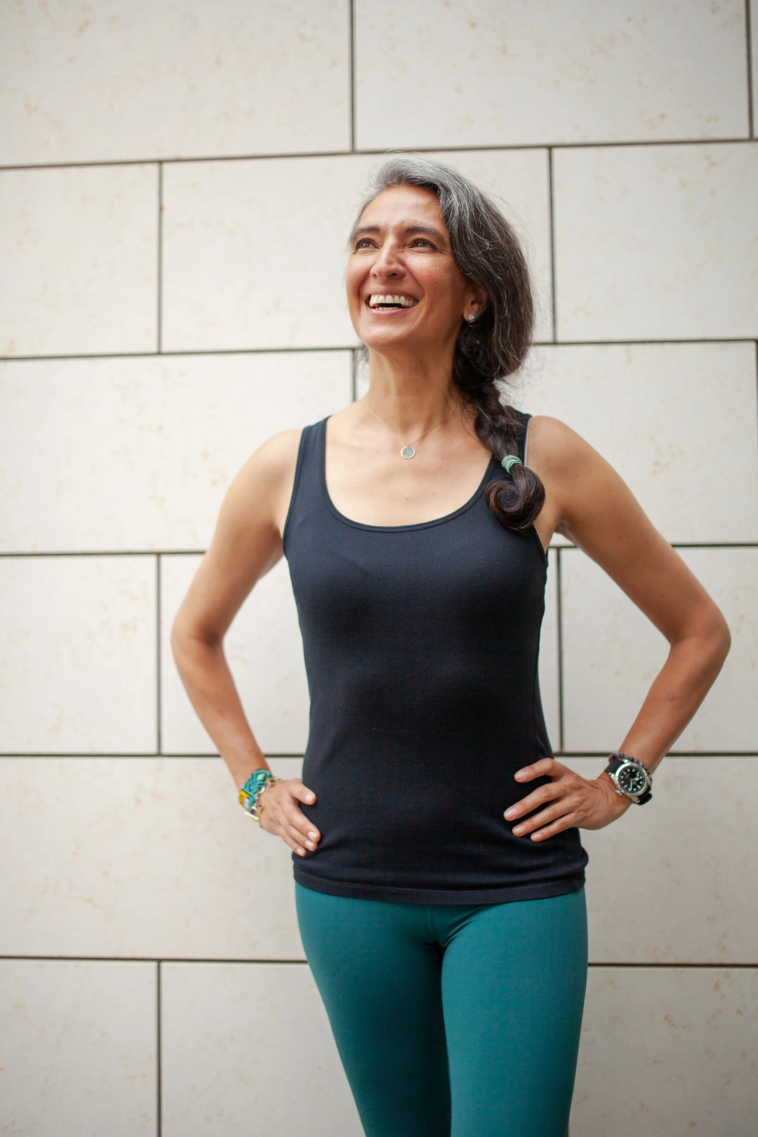 A smiling middle-aged woman with gray hair styled in a braid, wearing a black tank top and teal leggings, standing with hands on her hips in front of a tiled wall.