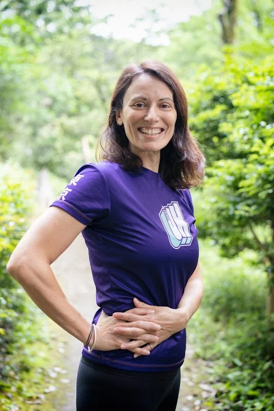 A woman with brown hair smiling and standing outdoors on a trail surrounded by green trees, wearing a purple t-shirt with a logo on the front and text on the sleeve.