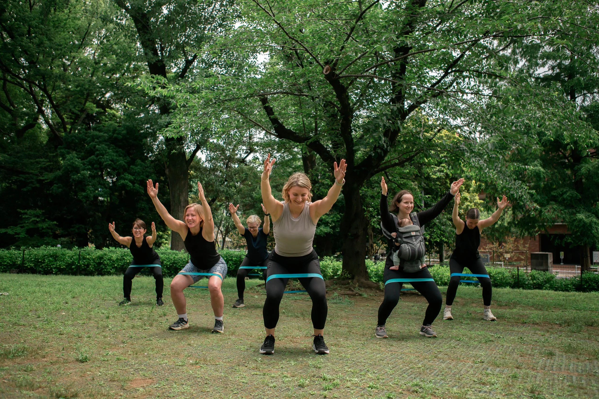 Group of women exercising outdoors in a park, wearing resistance bands around their thighs, with trees in the background.