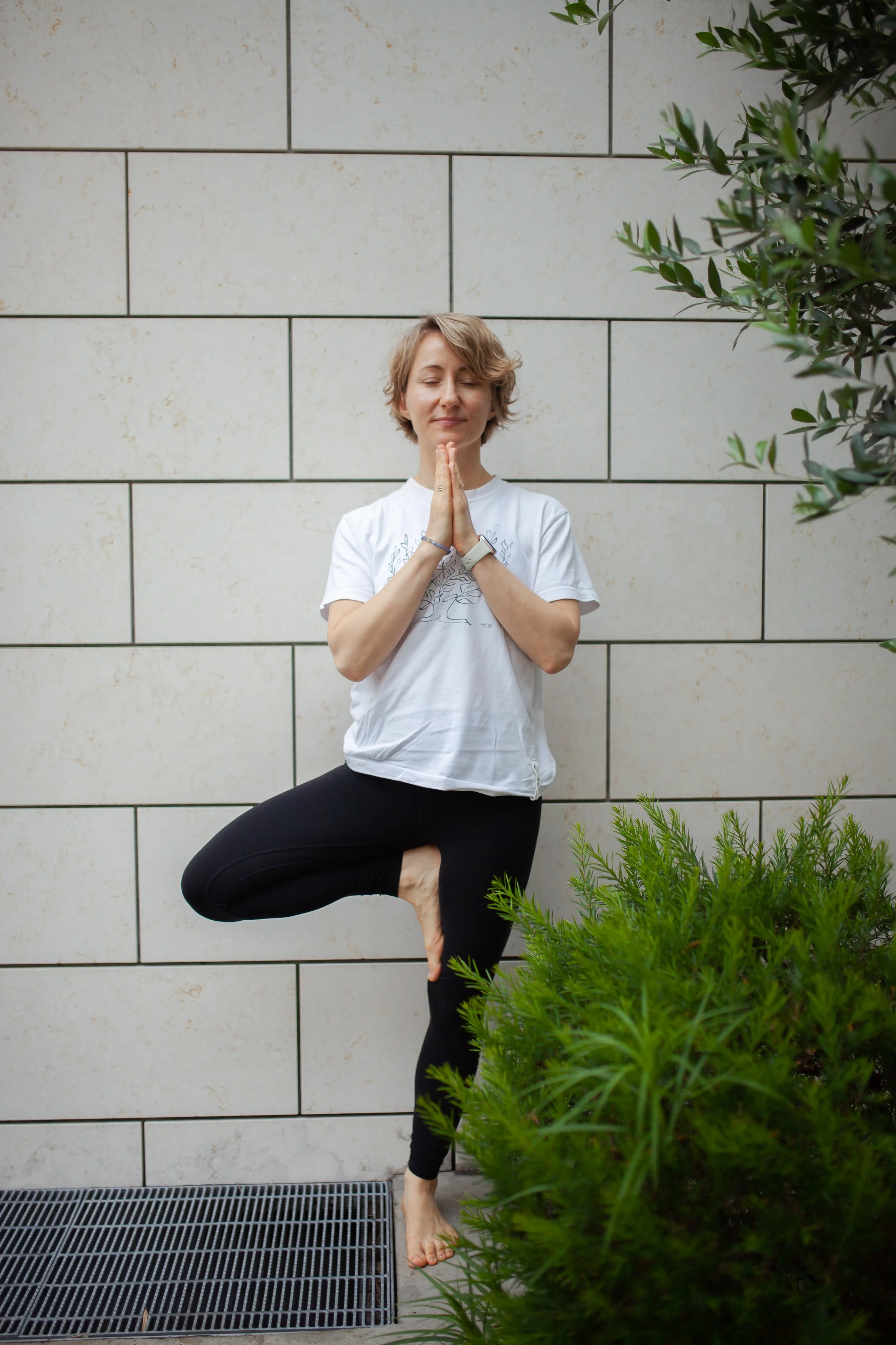 A woman practicing yoga outdoors in the tree pose, standing on one leg with her hands in a prayer position, next to green bushes and a tiled wall.