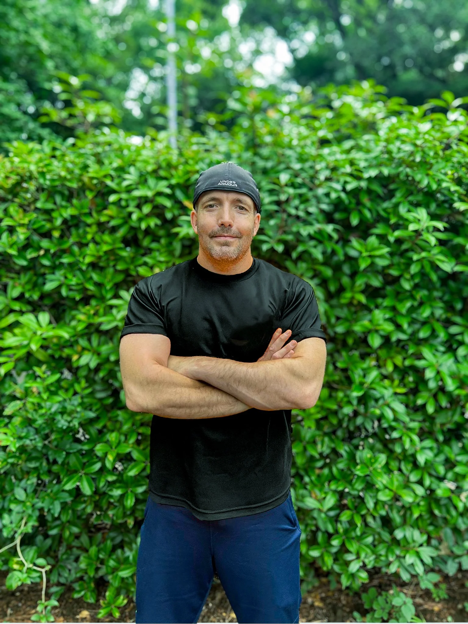 A man stands outdoors in front of a green hedge, crossing his arms and looking at the camera. He is wearing a black t-shirt, blue shorts, and a black cap.