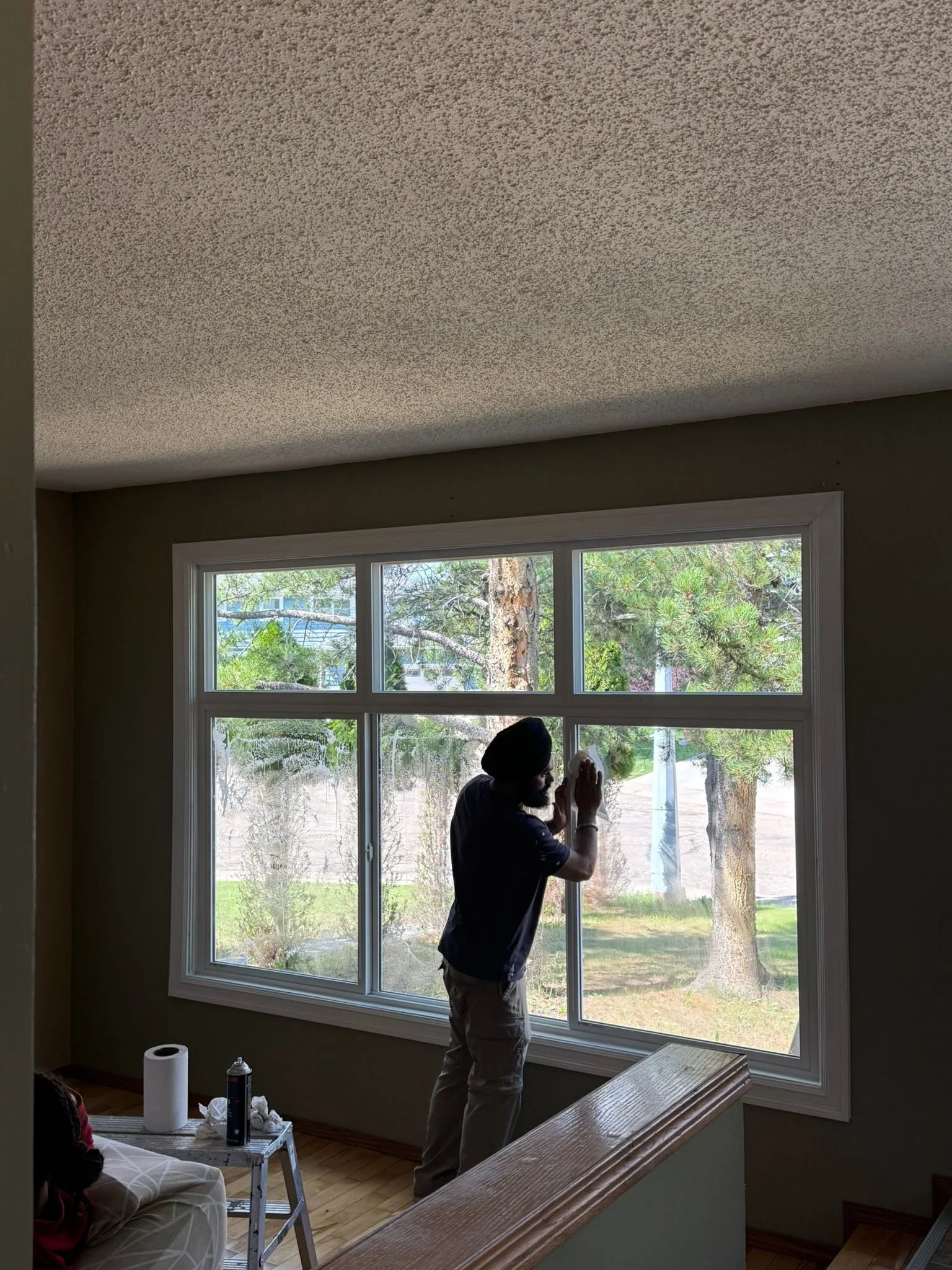 A man cleaning a large window in a living room during daytime, with trees visible outside.