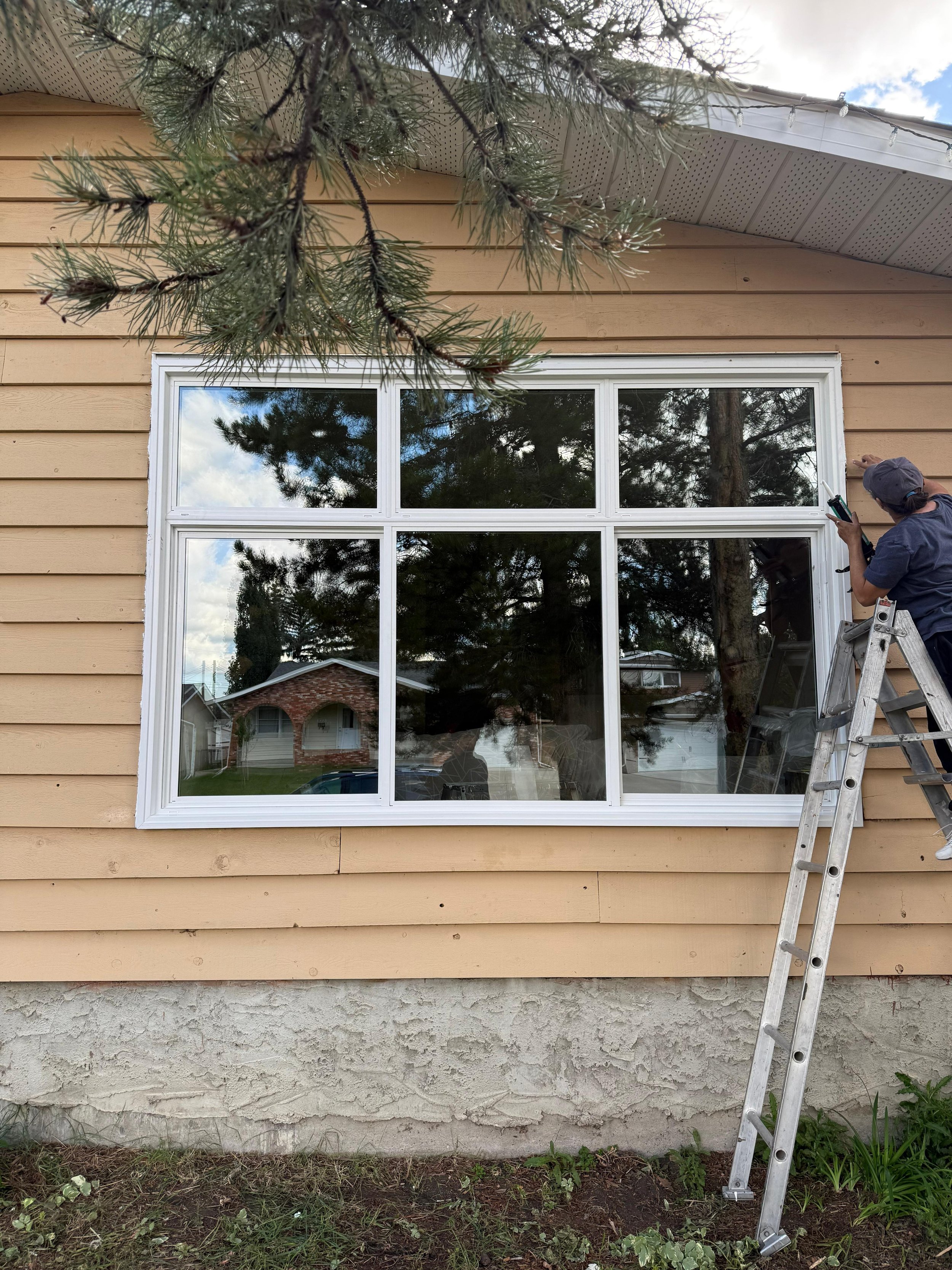 A person on a ladder installing or fixing a large window on the exterior of a house with tan siding.