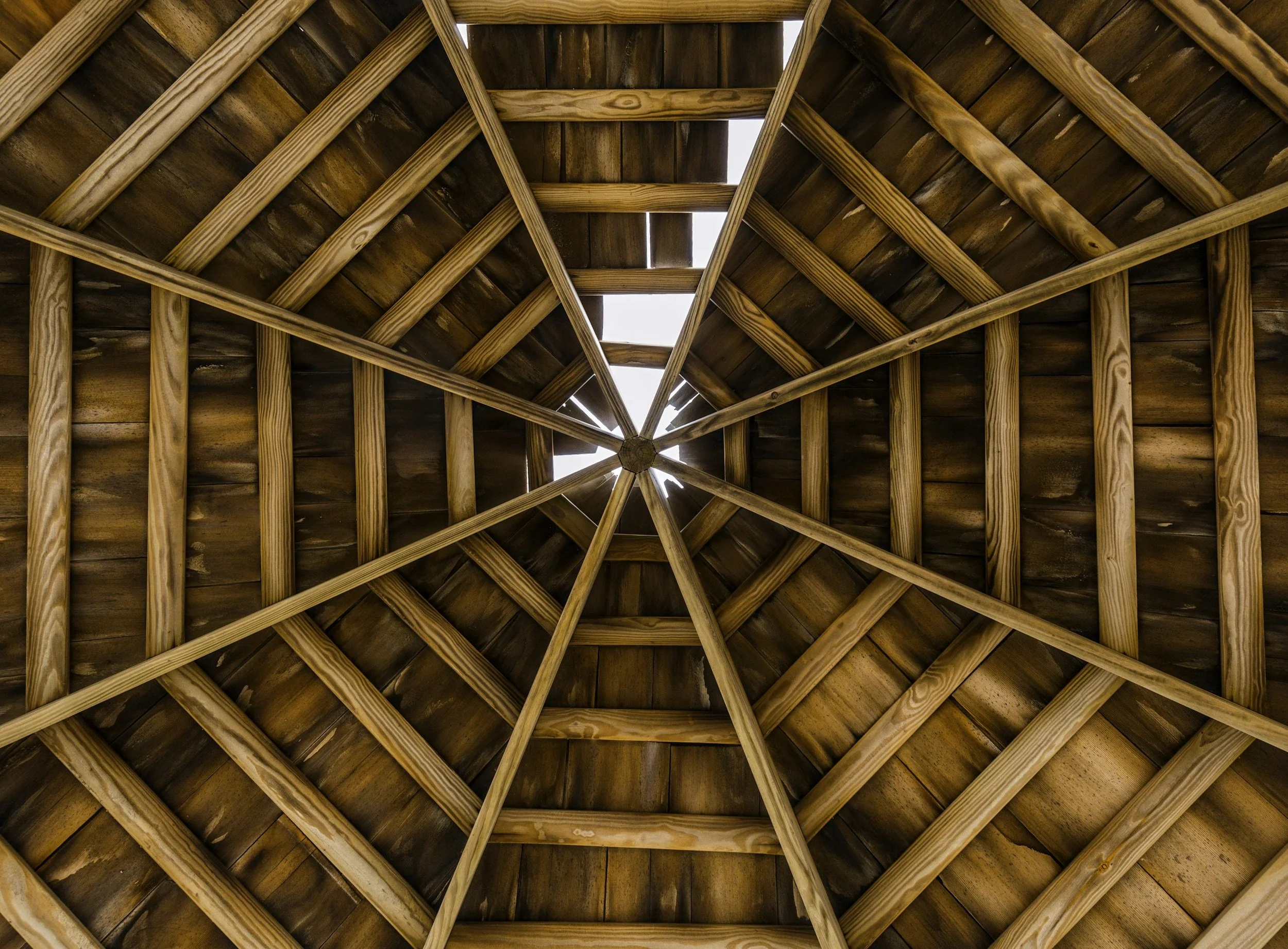Looking up at a wooden gazebo roof with a central hexagon, showing exposed beams and a small opening at the top with daylight shining through.