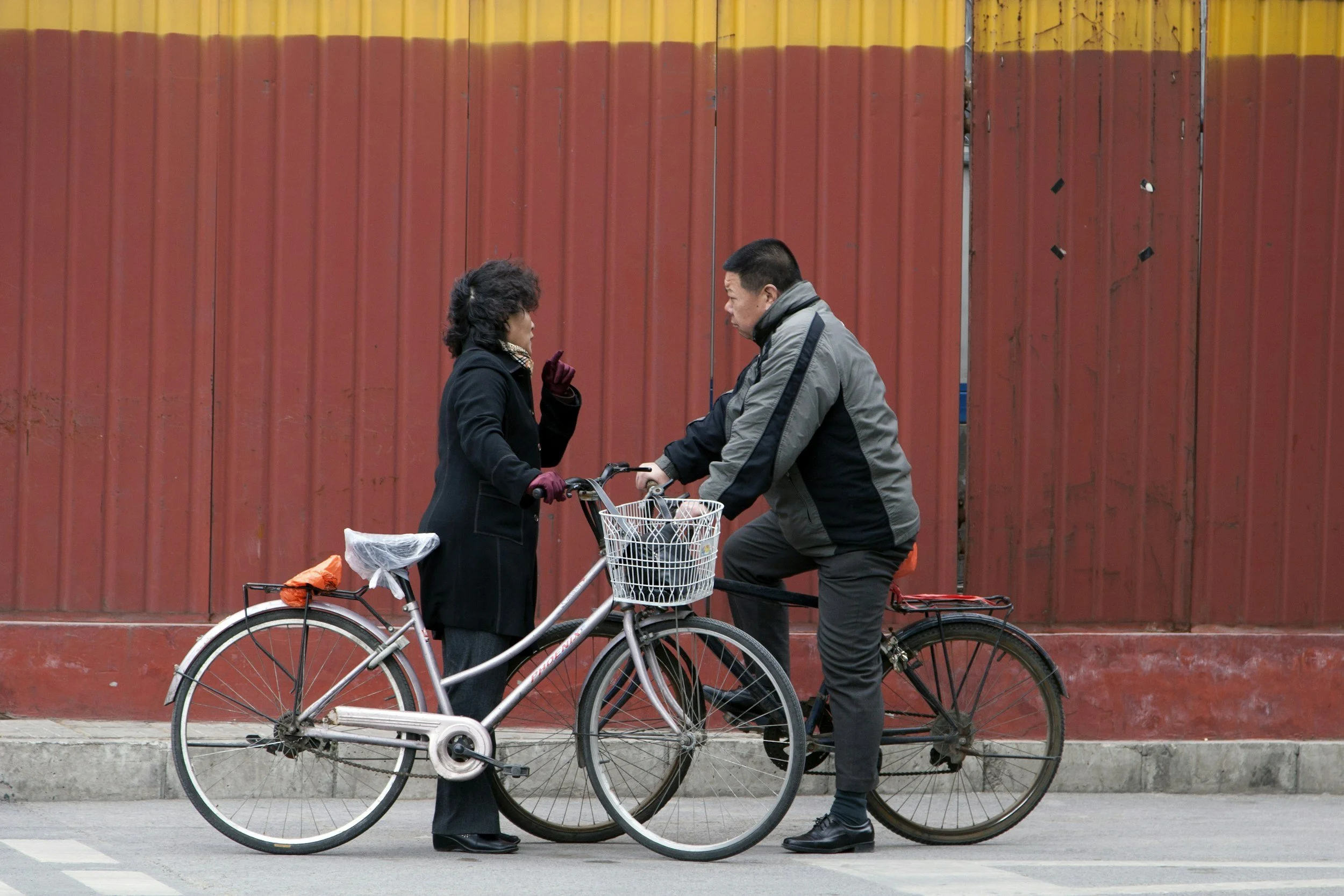 A man and a woman having a conversation beside their bicycles in front of a red wall. The woman appears to be talking with a finger raised, while the man listens with one foot on his bicycle pedal.