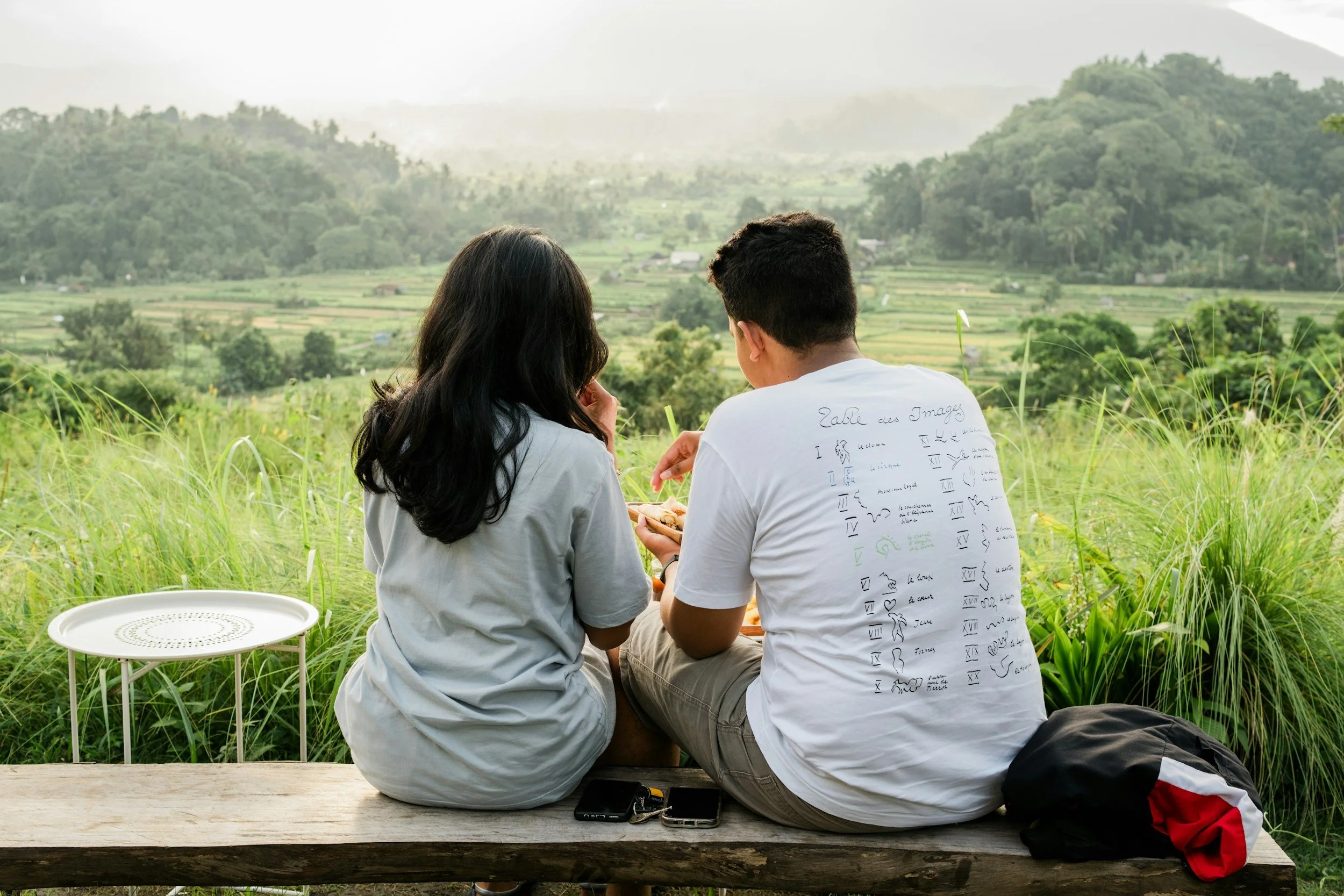 A man and woman sitting on a wooden bench overlooking a lush green landscape with terraced fields and mountains in the background.