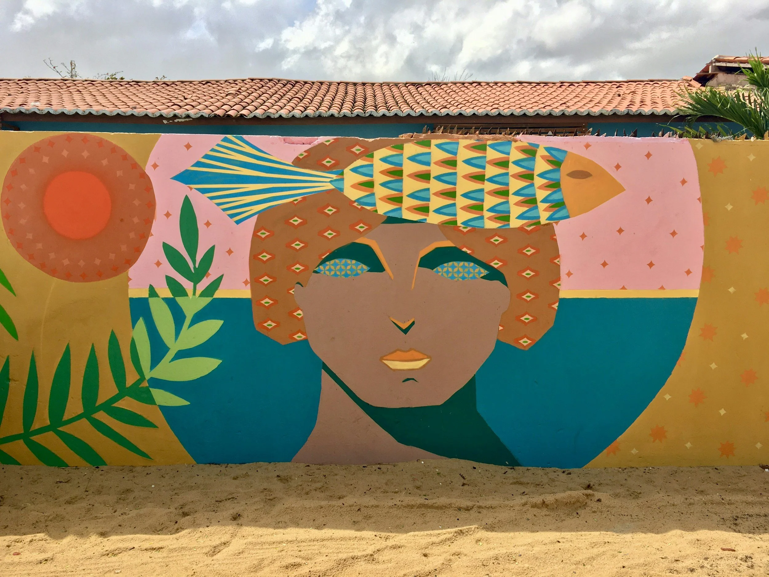 Colorful mural painting of a woman with a patterned headscarf, large earrings, and a tropical landscape, painted on a wall with a sky and rooftops visible above.