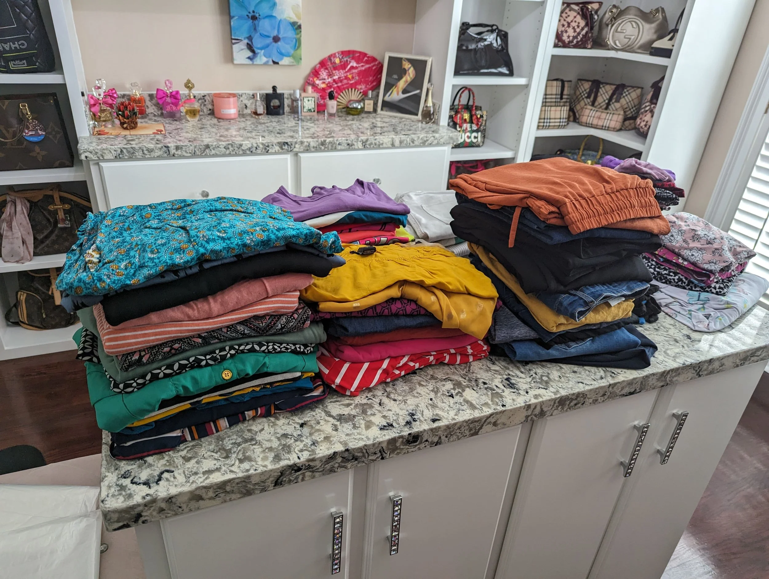Wash and fold laundry. Arranged stacks of colorful folded clothes on a kitchen countertop with a white cabinet in the background containing designer handbags and décor items.