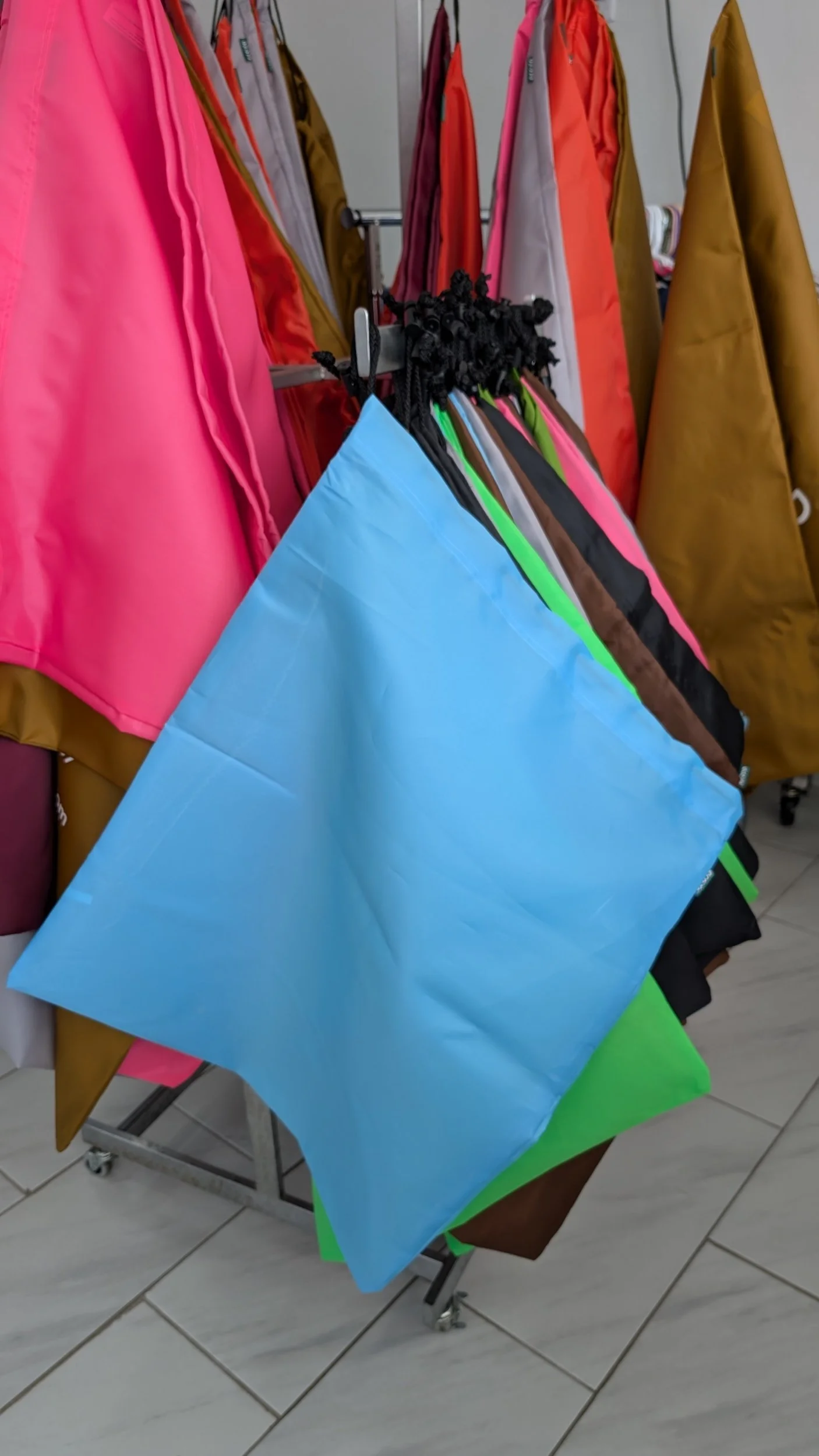 Medium flat rate bags for full service pick up and delivery. Colorful nylon drawstring bags hanging on a rack, including blue, green, pink, black, and brown, with jackets in various colors hanging in the background.