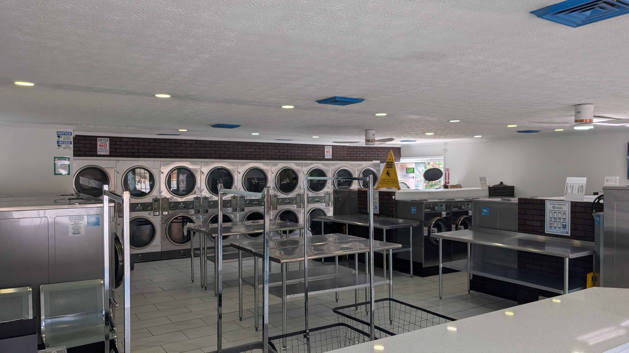 Bright WSHFLD laundromat interior with front-loading washers and dryers, stainless-steel folding tables, tiled floors, and ceiling lights.