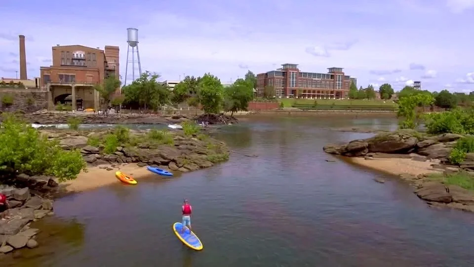 Columbus, Georgia, Riverfront, People kayaking on a river with rocks, trees, and buildings in the background.