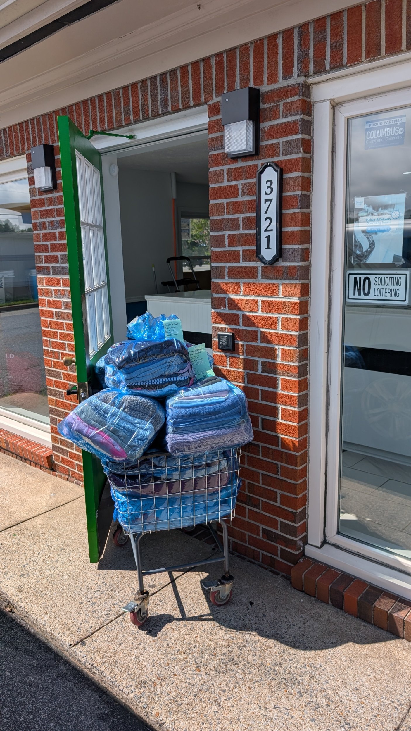 A Laundry cart outside Wshfld Laundry in Columbus GA, is filled with folded blankets and a large plastic bag of water bottles outside a brick building with a glass door showing a sign that says 'No Soliciting Loitering.'