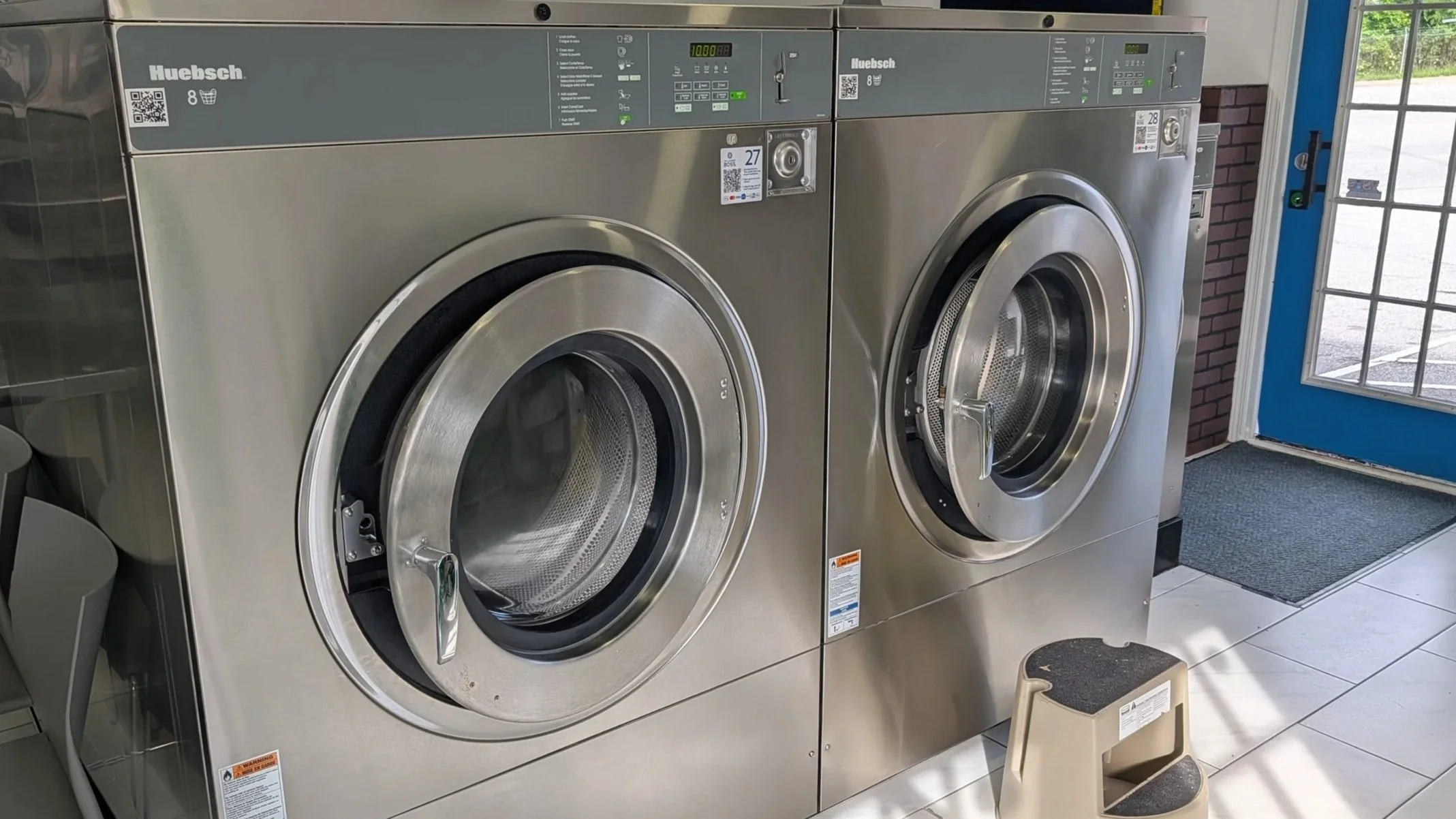 Two large-capacity 8-load commercial front-loading washing machines inside a modern self-service laundromat. The stainless steel washers are positioned side by side with digital control panels and oversized glass doors, designed to handle bulky items