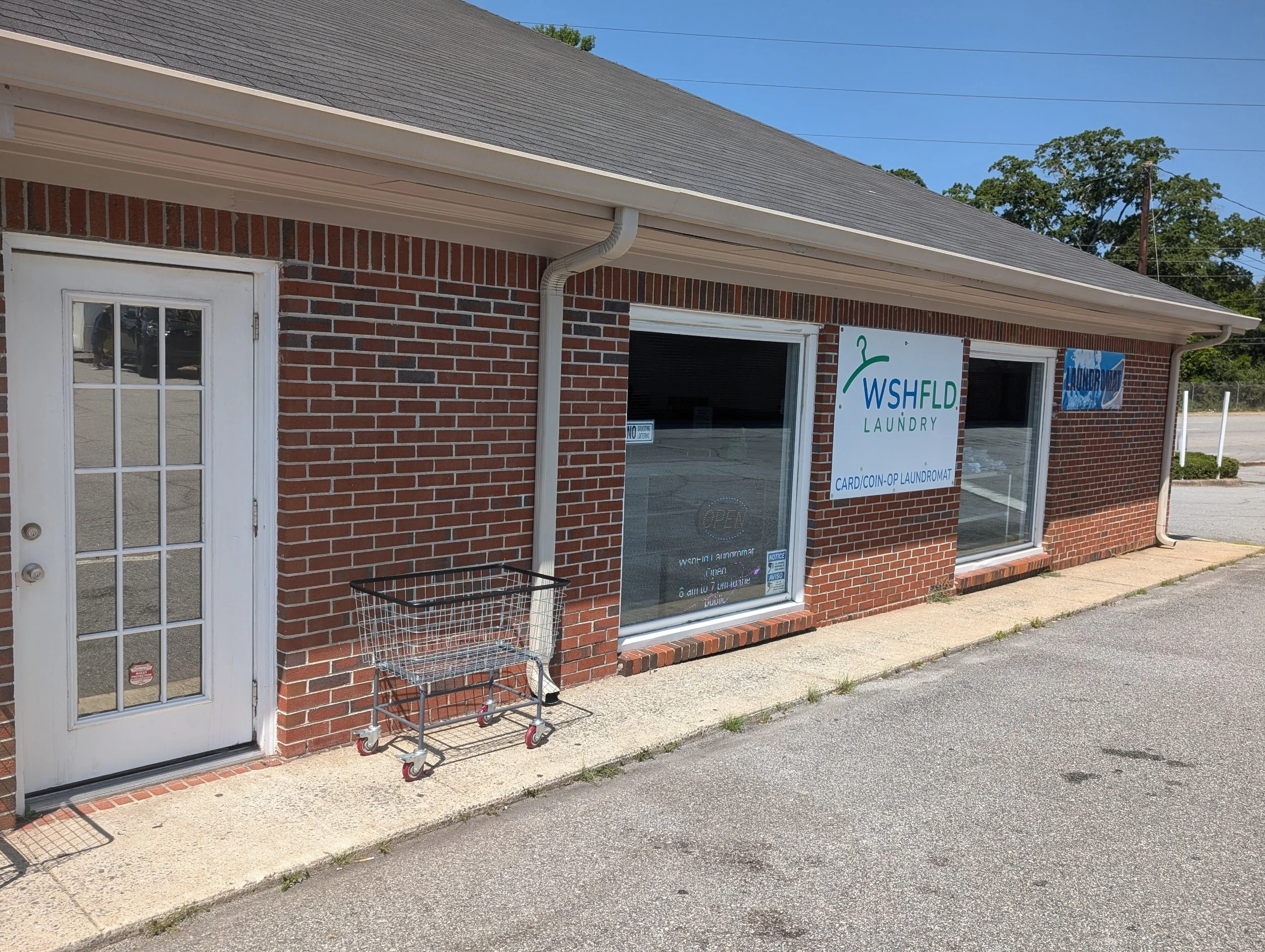 Entrance to wshfld laundromat. Brick building with signs for Wash Fold Laundry and Coin Laundry, a shopping cart outside, and a glass door.