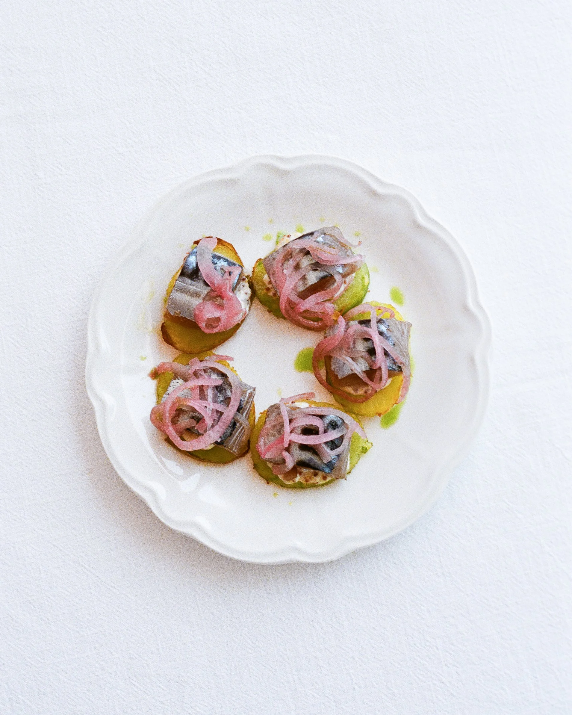 Plate with six small servings of sliced potatoes topped with sardines and pink pickled onions, garnished with green sauce, on a white scalloped edge plate.