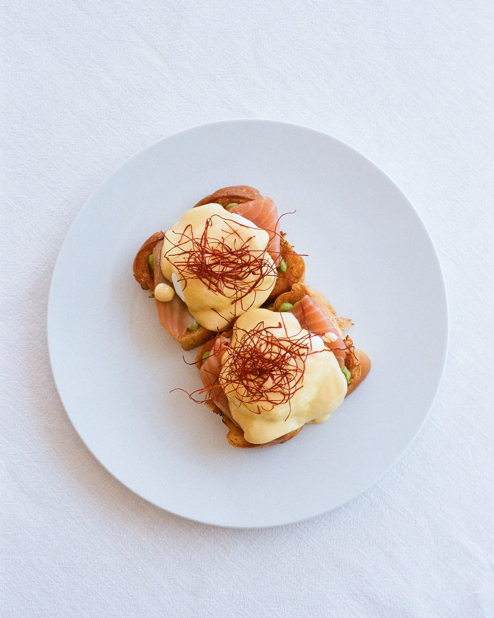 Two pieces of eggs Benedict with smoked salmon, hollandaise sauce, and red chili threads served on a white plate.