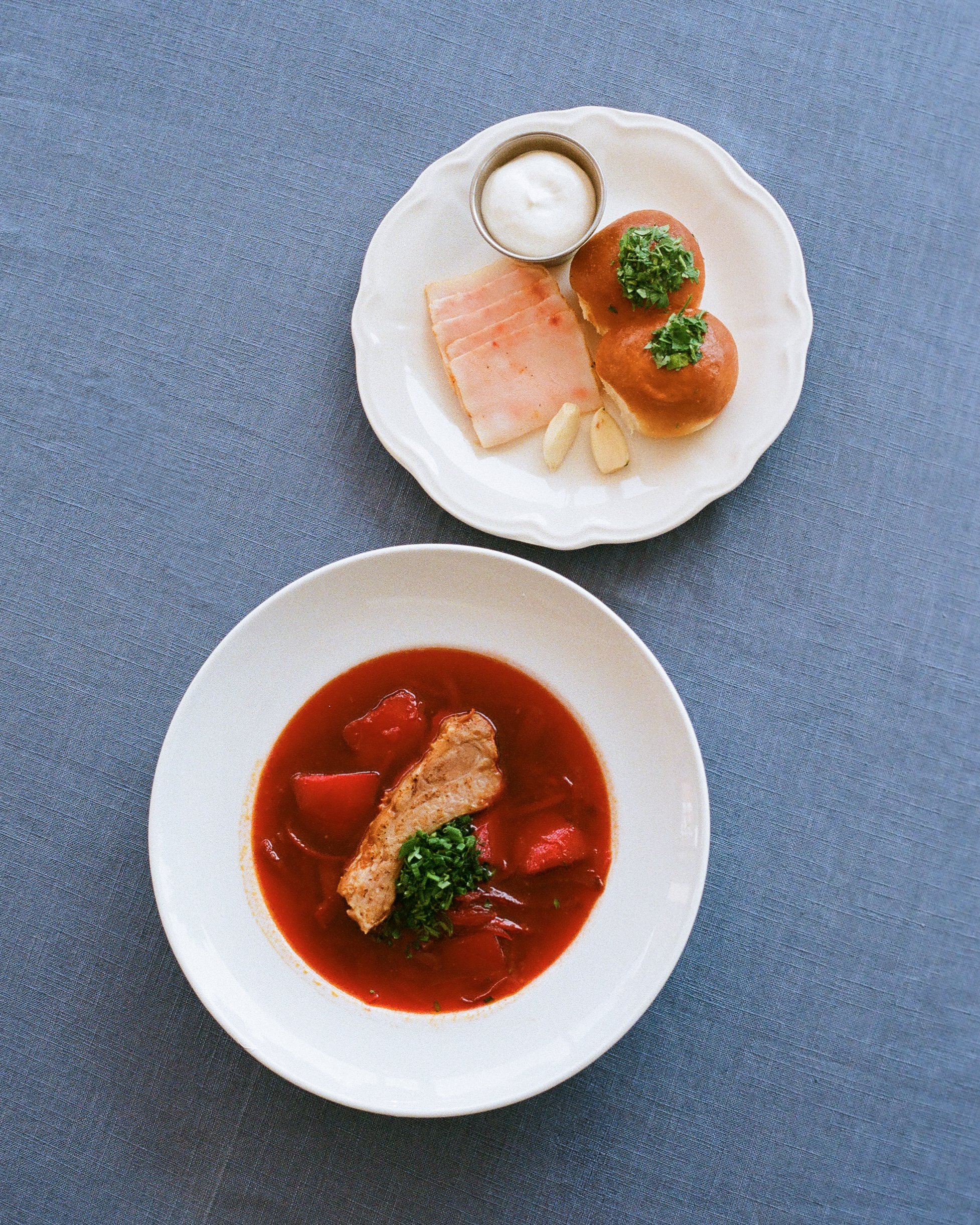 A top view of two white plates on a gray table. The upper plate has two bread rolls topped with parsley, a small cup of white sauce, slices of ham, and garlic cloves. The lower plate contains a bowl of tomato soup with a piece of chicken, tomatoes, a
