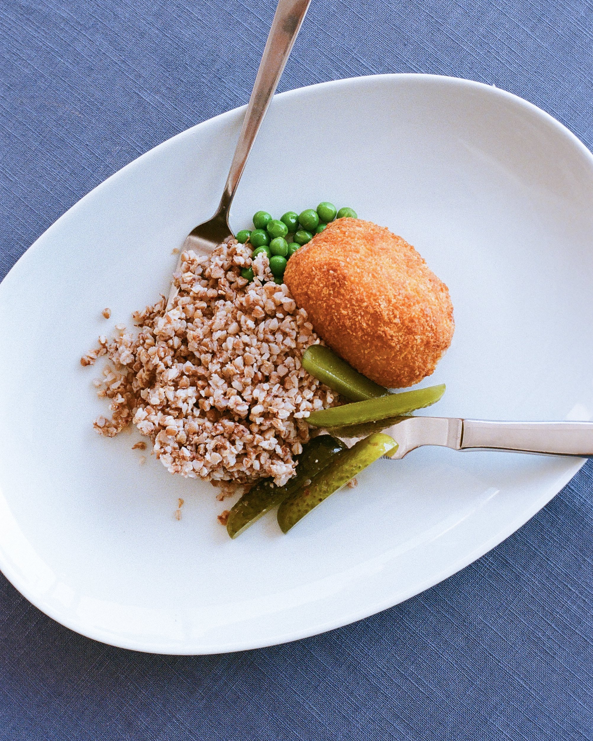 A white plate with cooked ground meat, green peas, pickles, and a breaded fried item, garnished with a fork and knife, on a dark blue tablecloth.