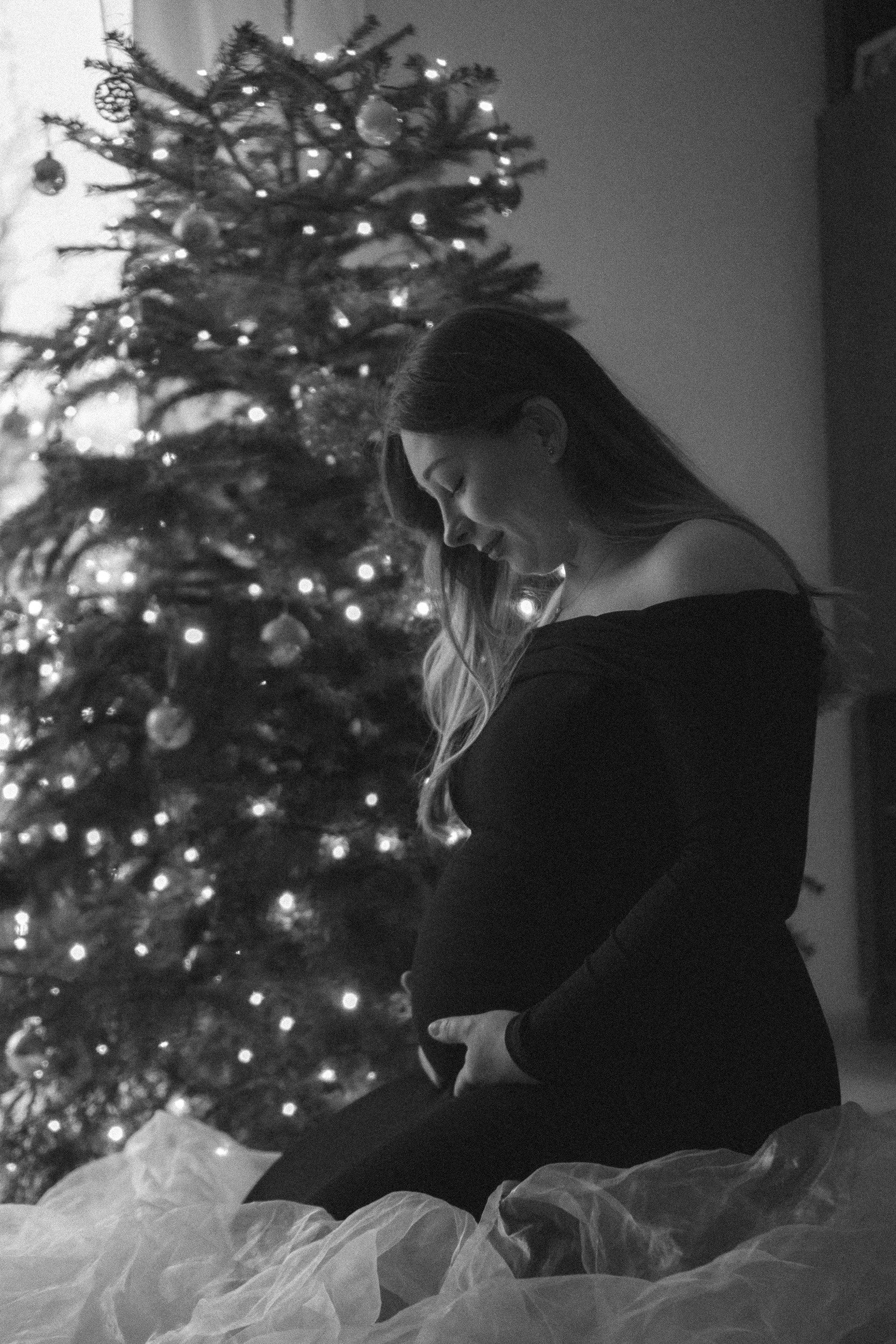 Schwangere Frau sitzt vor einem geschmückten Weihnachtsbaum, lächelt und hält ihren Bauch, schwarz-weiße Fotografie.