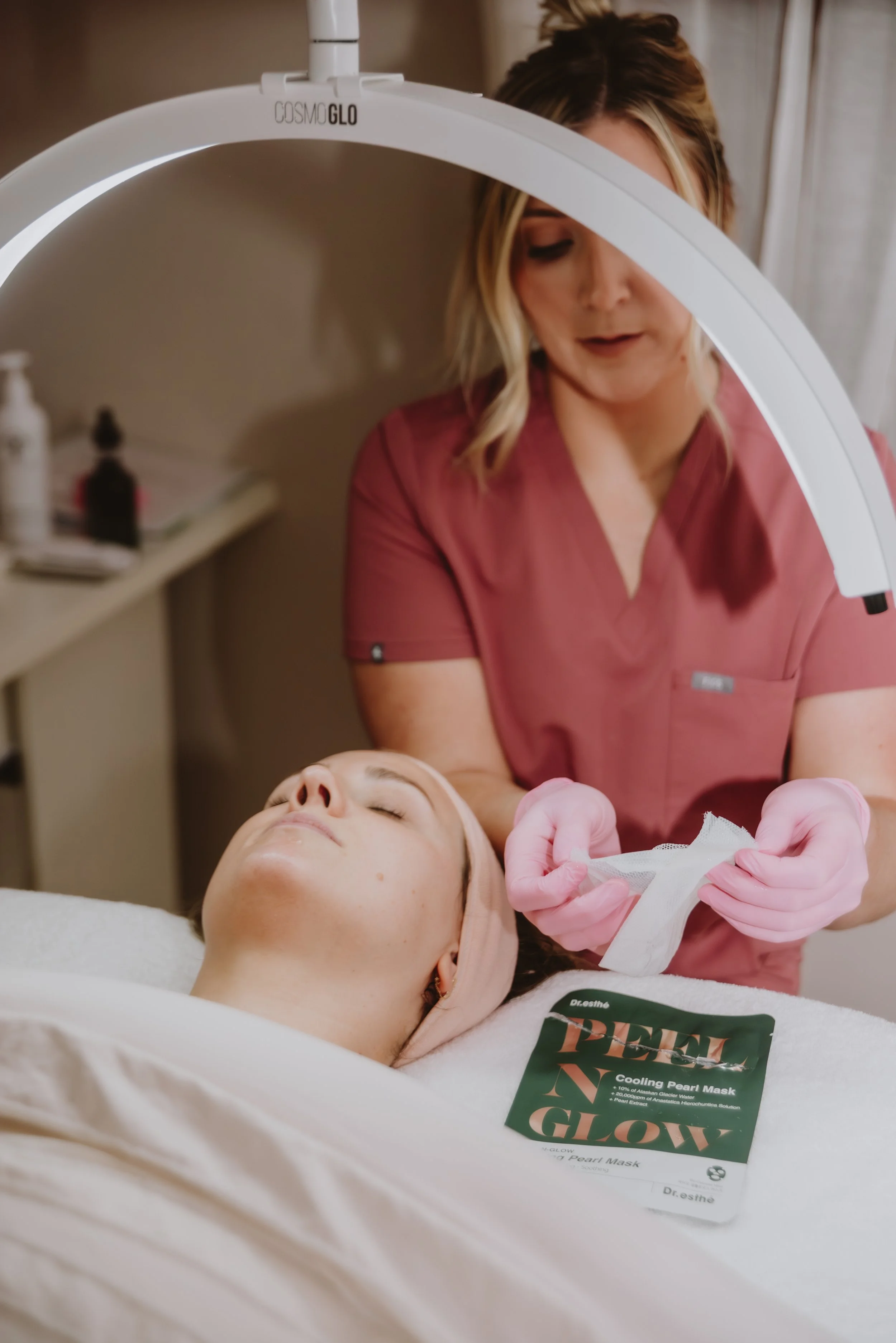 A woman lying on a treatment bed with her eyes closed while a skincare professional applies a facial mask. The professional is wearing pink gloves and a pink uniform, and is working under a circular LED light. There is a green facial mask packet labeled 'PEEL GLOW' on the bed next to the woman.