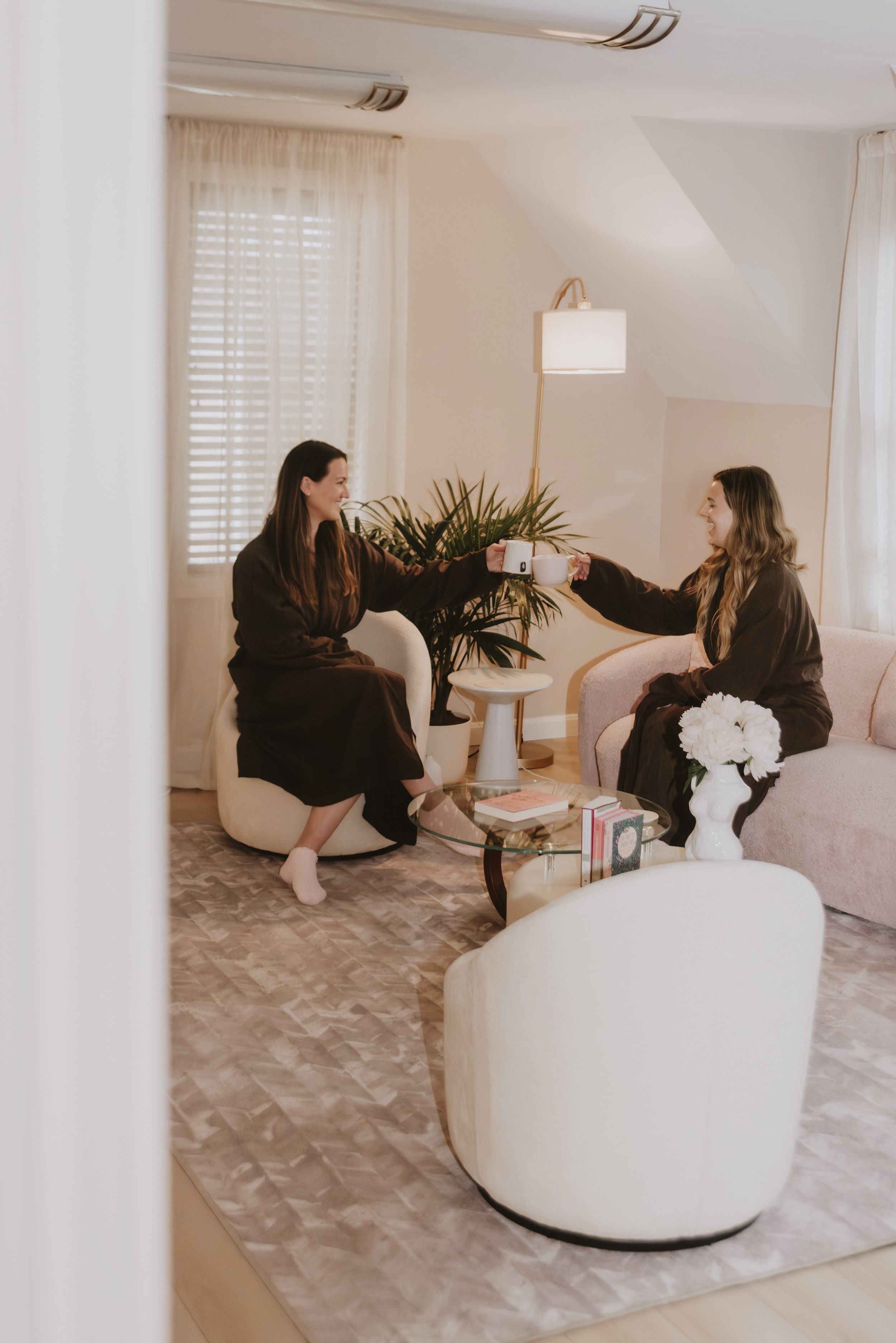 Two women are sitting in a cozy, well-lit living room, clinking mugs in a friendly toast. The room features cream-colored furniture, a glass coffee table with books, a pink sofa, a white armchair, and a large potted plant.