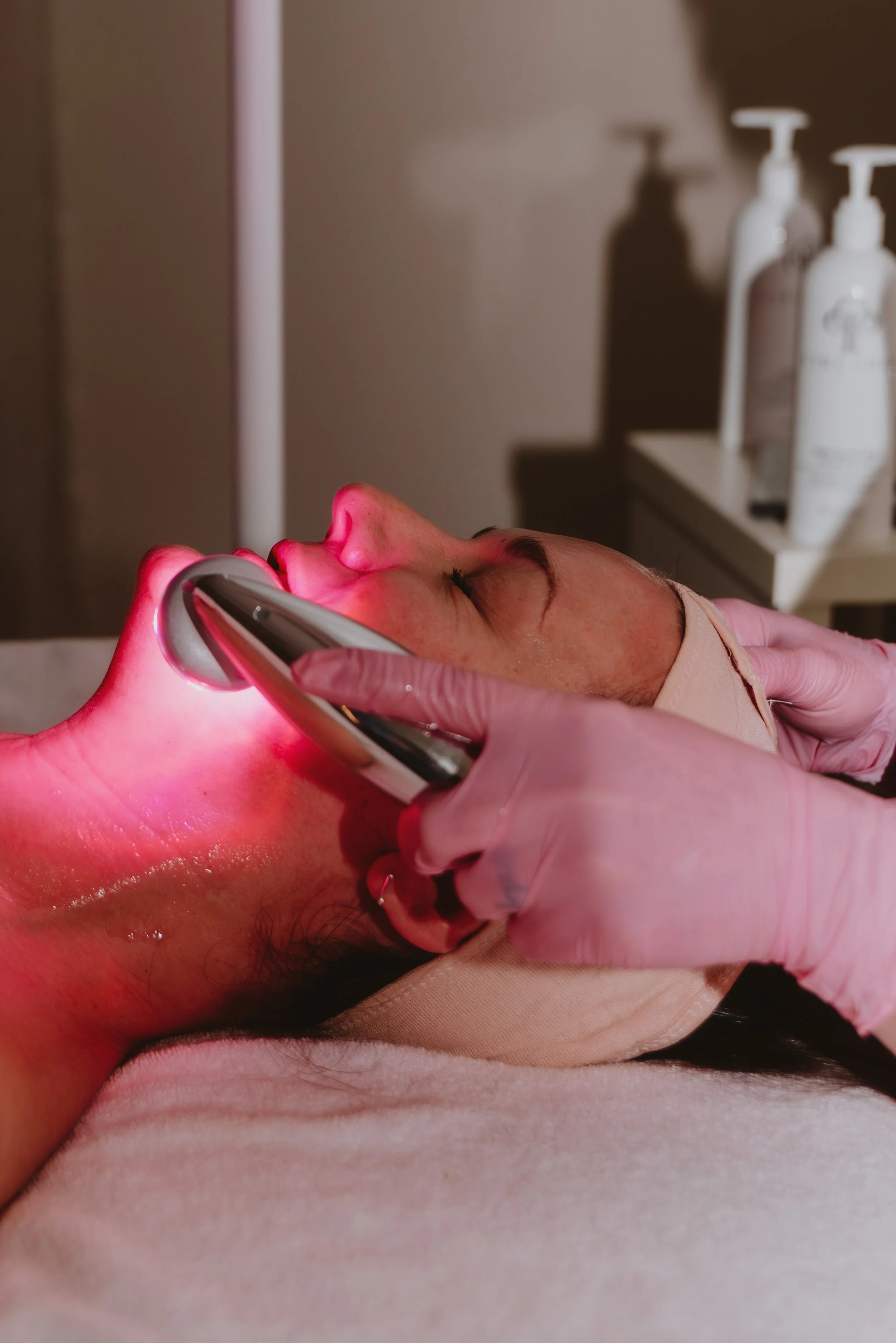 A woman receives a facial treatment with a red LED light, lying on a treatment bed with her eyes closed, while a practitioner in pink gloves holds the device against her face.