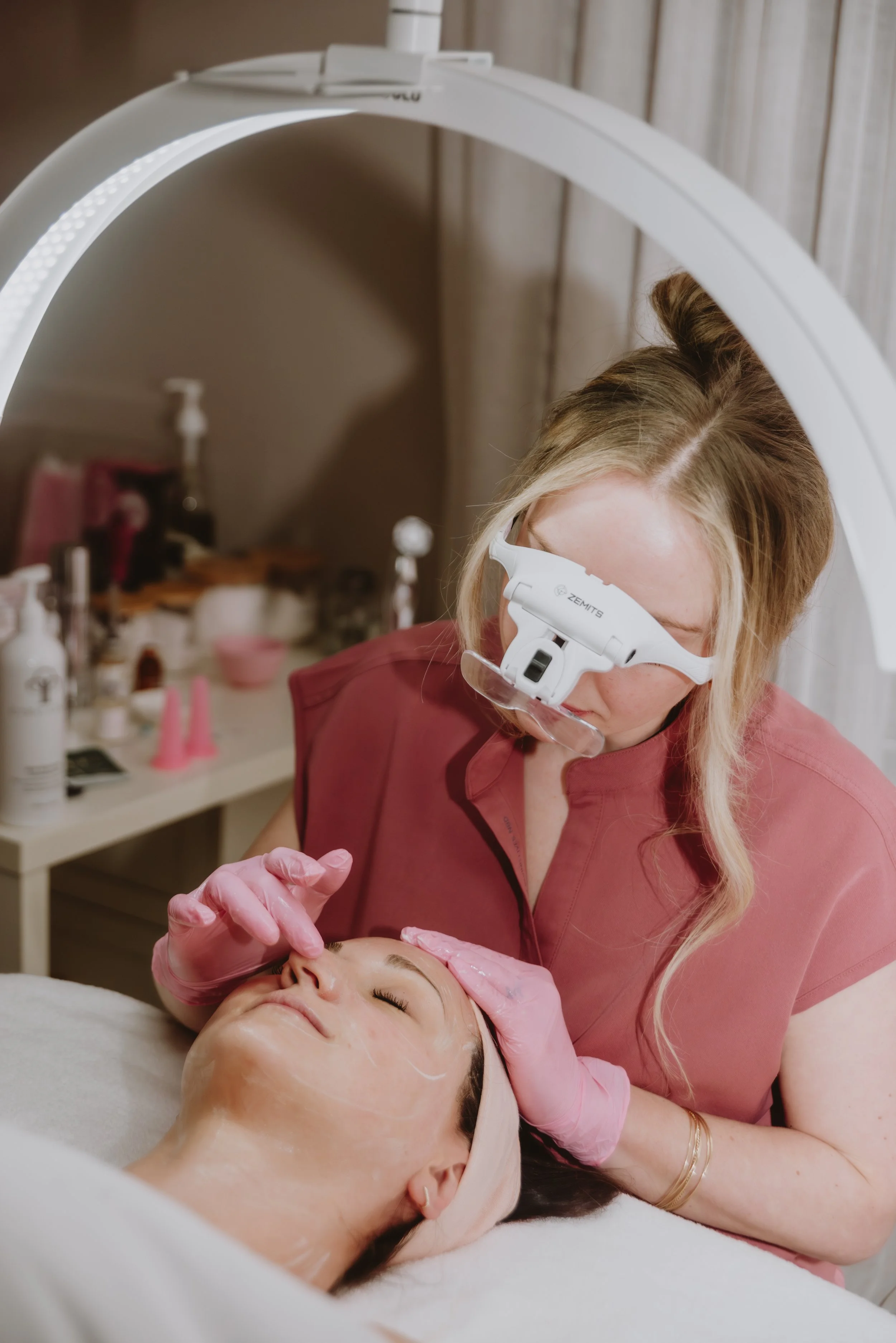 A woman lying on a treatment bed receiving a facial treatment from a skincare professional using a magnifying headset.