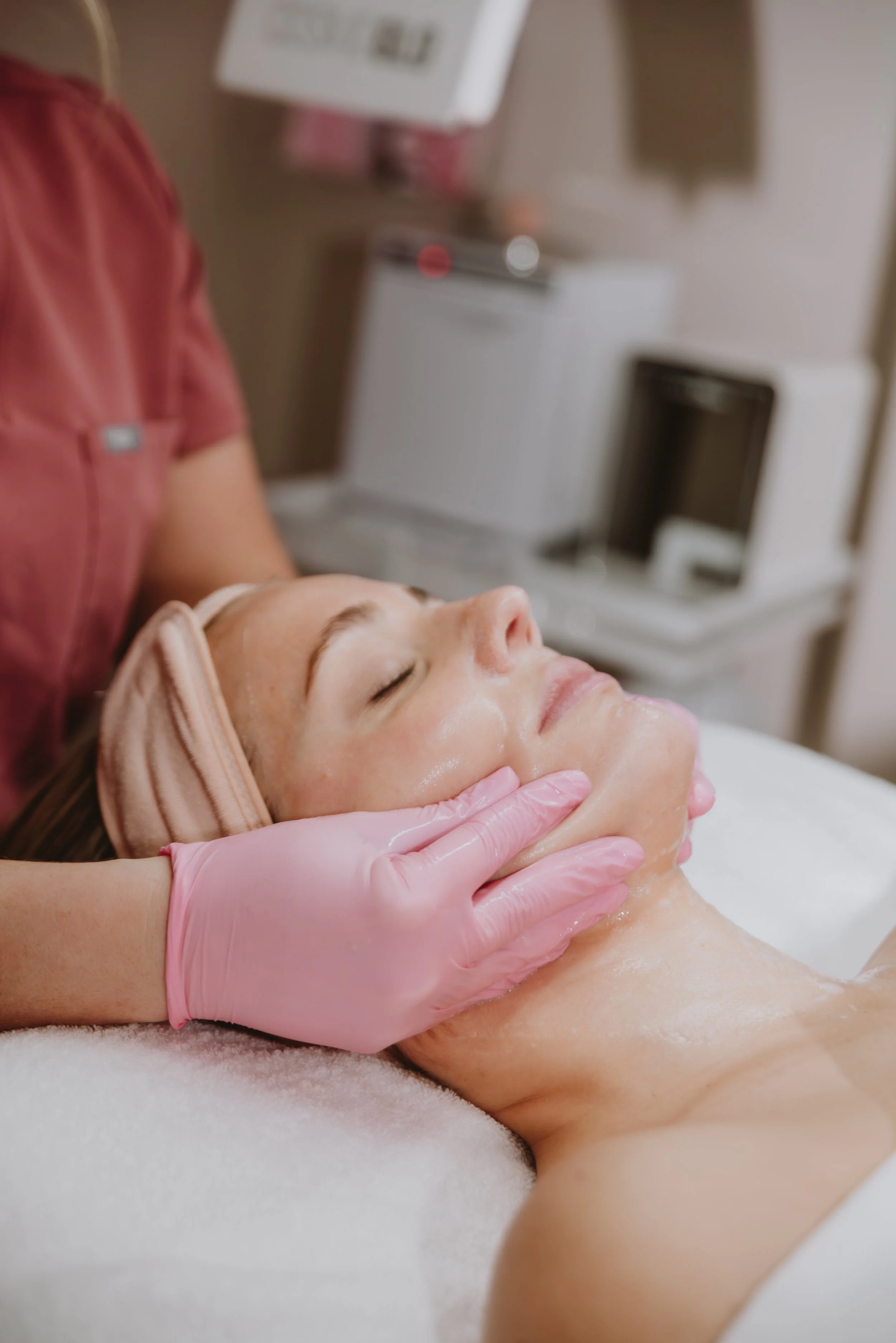 A woman receiving a facial treatment at a spa, lying down with eyes closed, wearing a headband, with a skincare professional wearing pink gloves massaging her face.