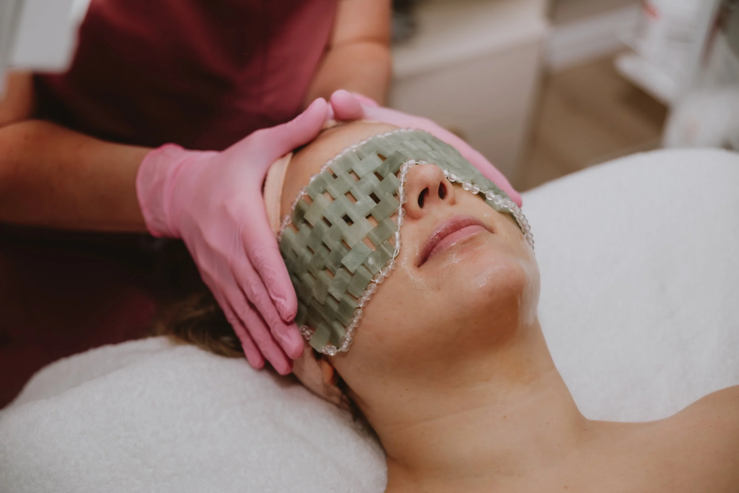 A woman lying down during a facial treatment with a green eye mask and a lip mask, while a practitioner wearing pink gloves gently holds her forehead.