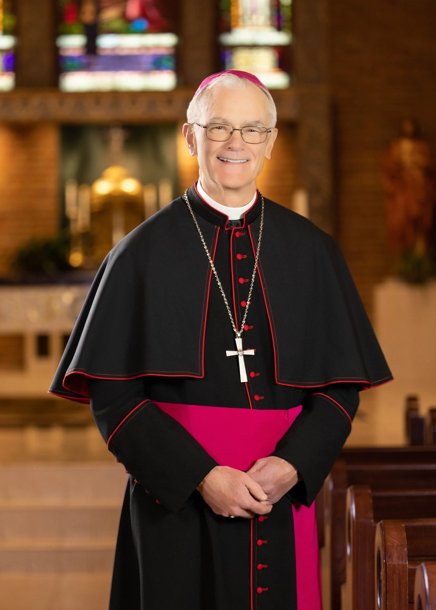 A man wearing a purple religious robe with a large cross necklace and a purple skullcap, standing outdoors against a stone building.