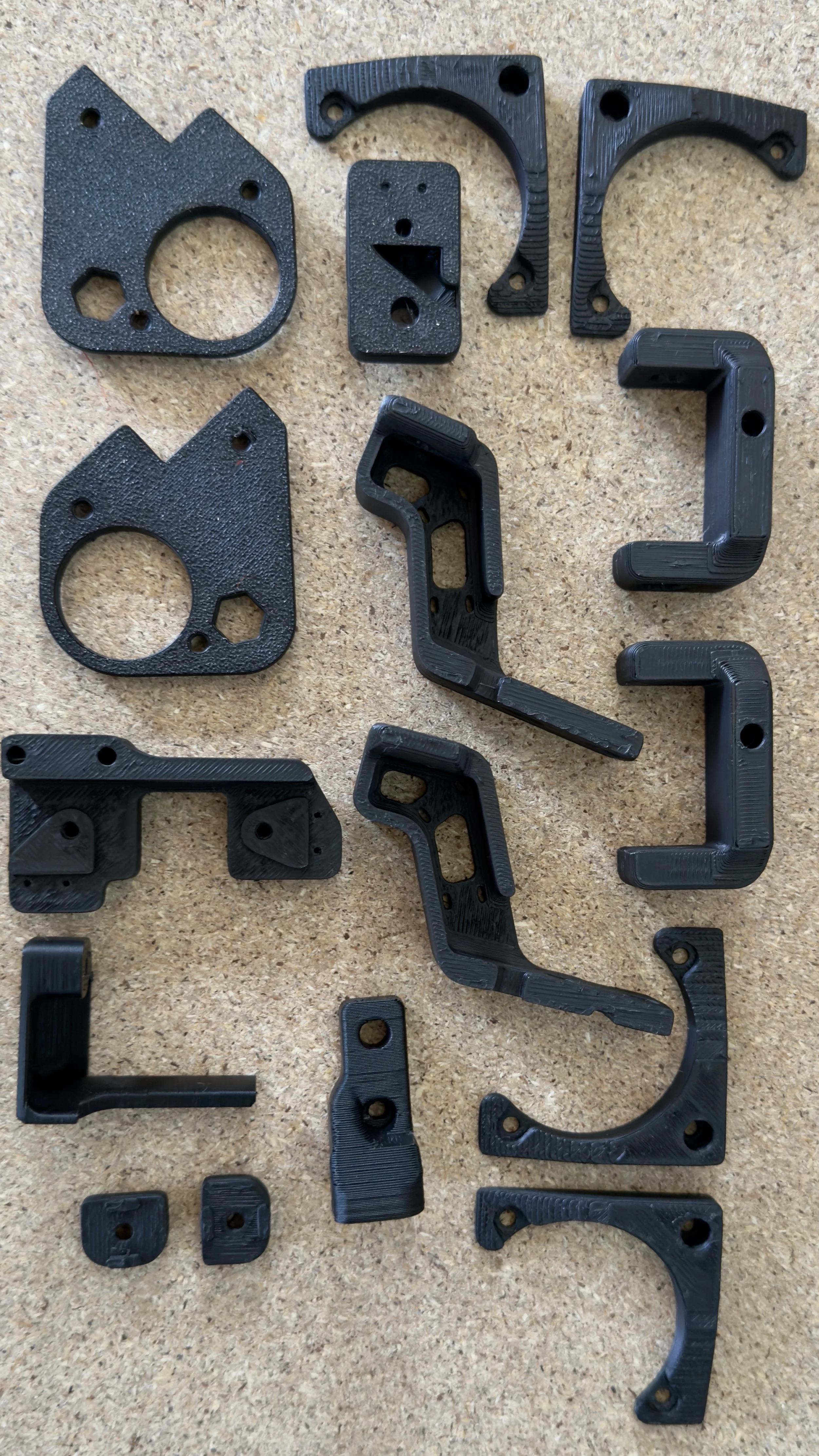 Set of black 3D-printed mechanical parts on a cork surface, including brackets, mounting plates, and hooks.