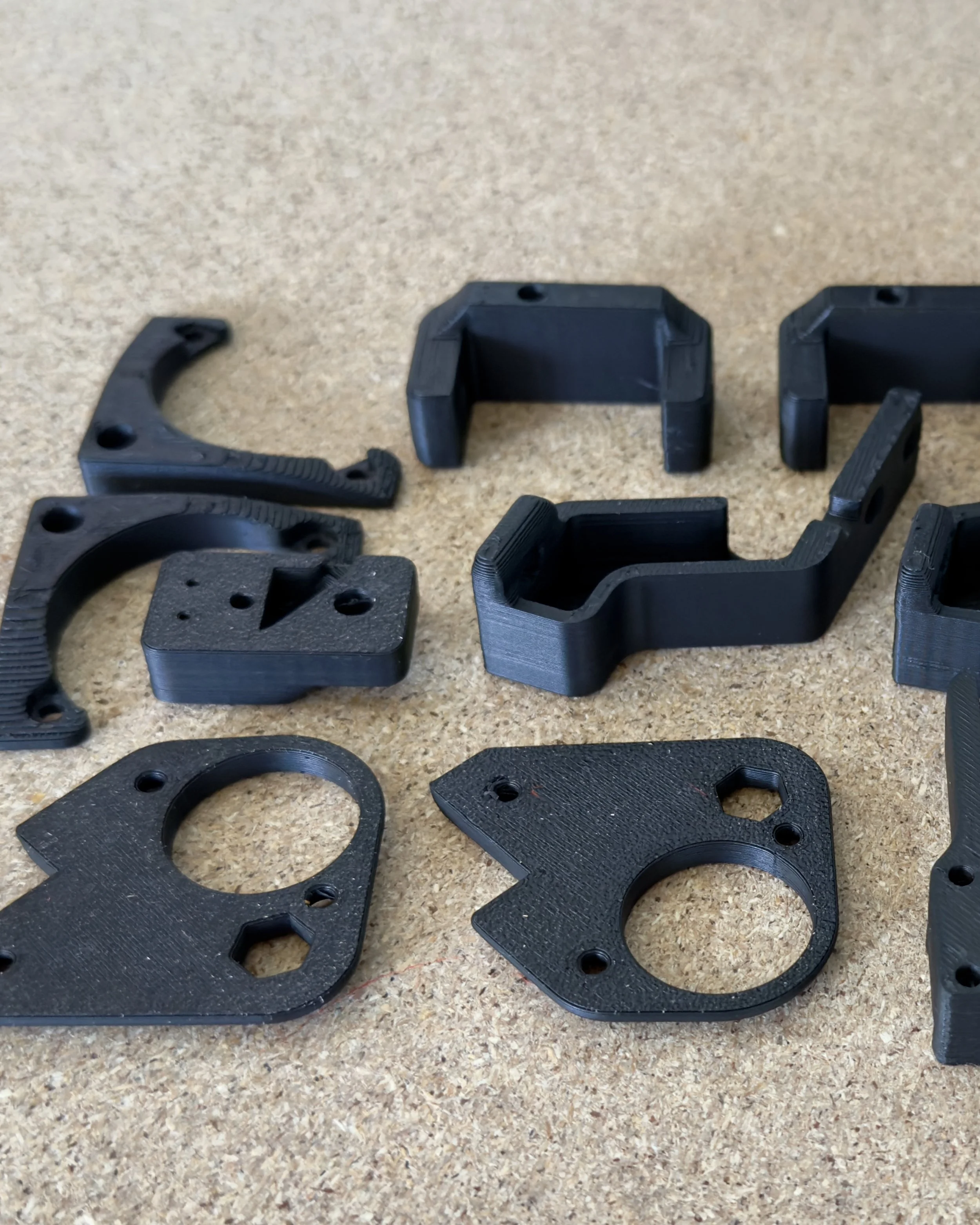 Various black 3D-printed plastic parts with different shapes and sizes laid out on a wooden surface.