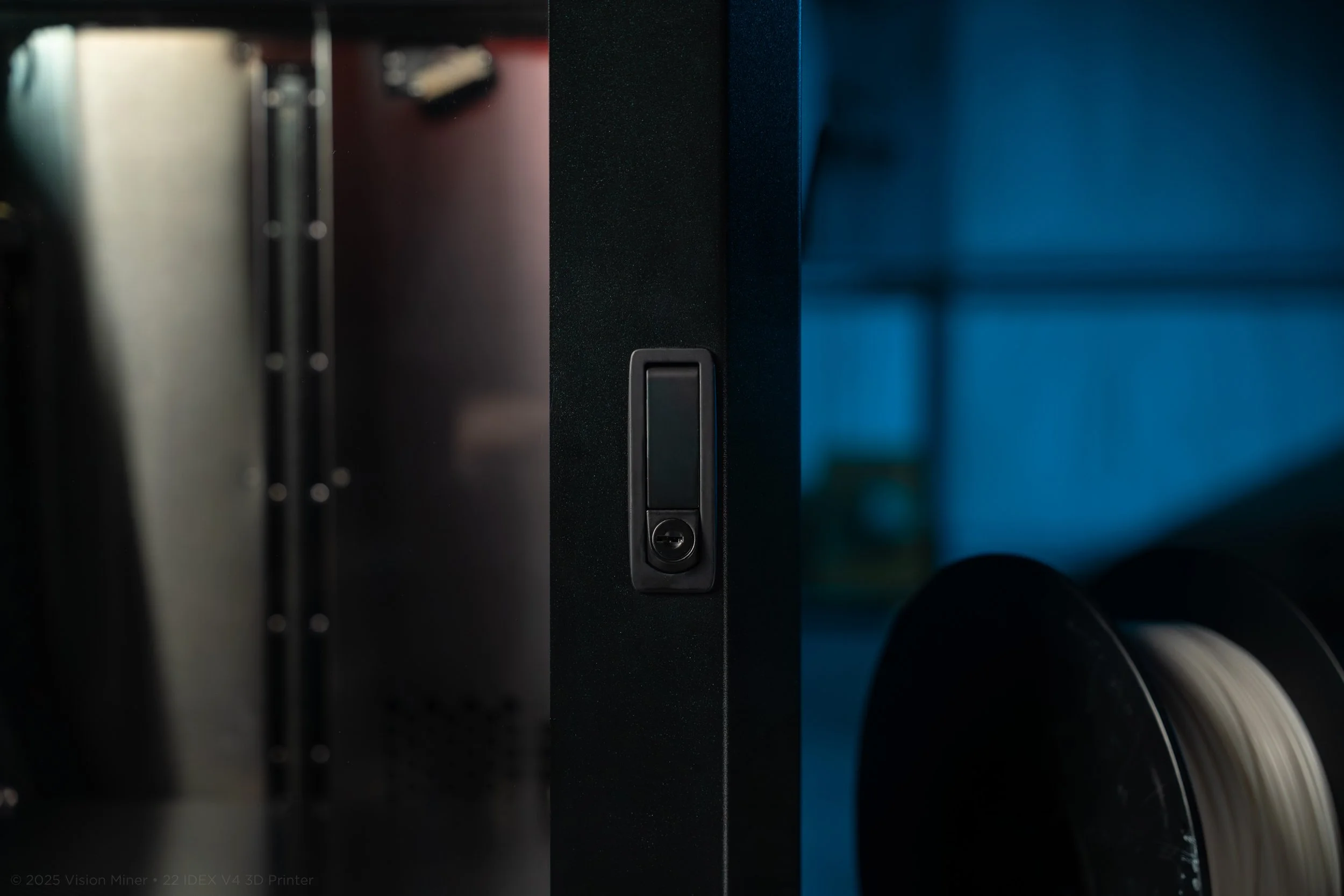 Close-up of a black 3D printer showing the front panel with a vertical slot and a circular button, with colored spools of filament visible on the right and black metal components on the left.