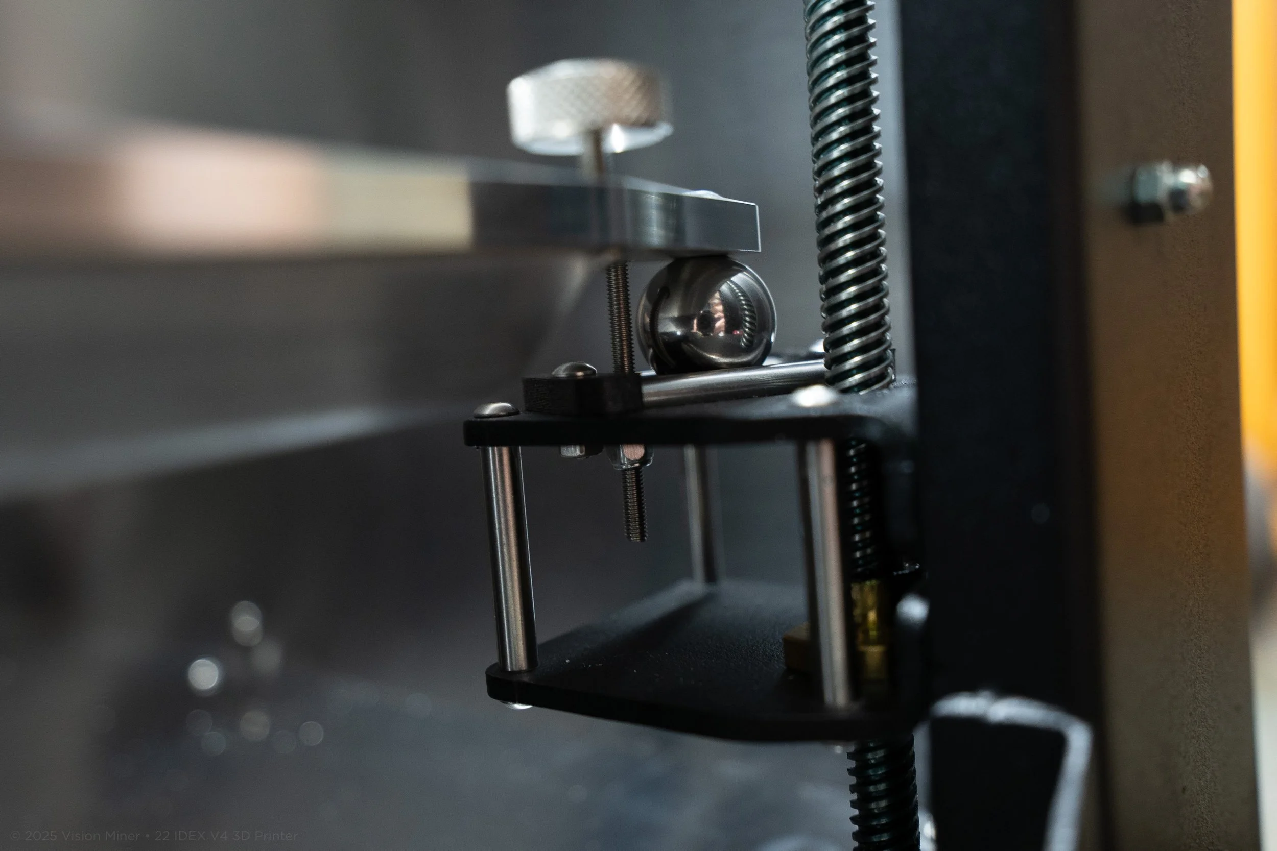Close-up of a metal mechanical part with a shiny steel ball, mounted on a black fixture with supporting rods and a large threaded screw, part of a machine or industrial equipment.
