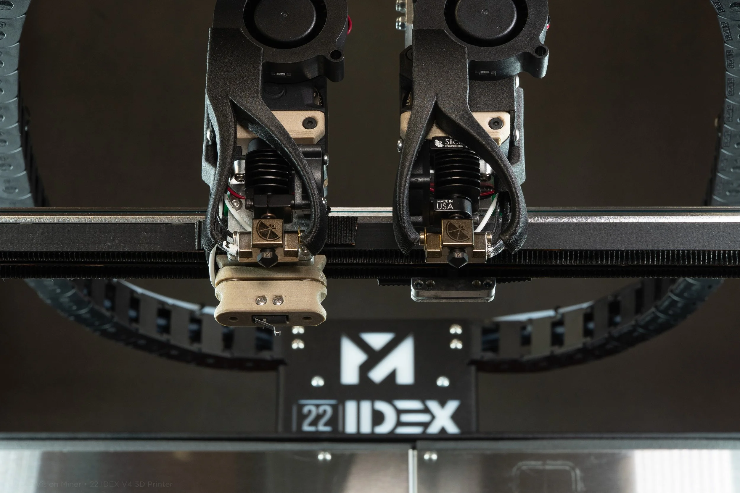 Close-up of a 3D printer's moving components, including the print head and an X-axis rail, with the logo of IDEX (Independent Dual Extrusion) visible at the bottom.