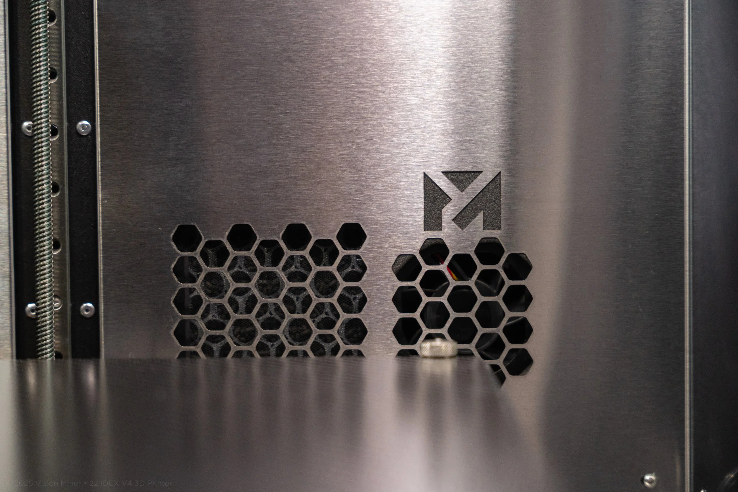 Close-up of a metallic 3D printer with hexagonal ventilation holes and a logo on the right side.