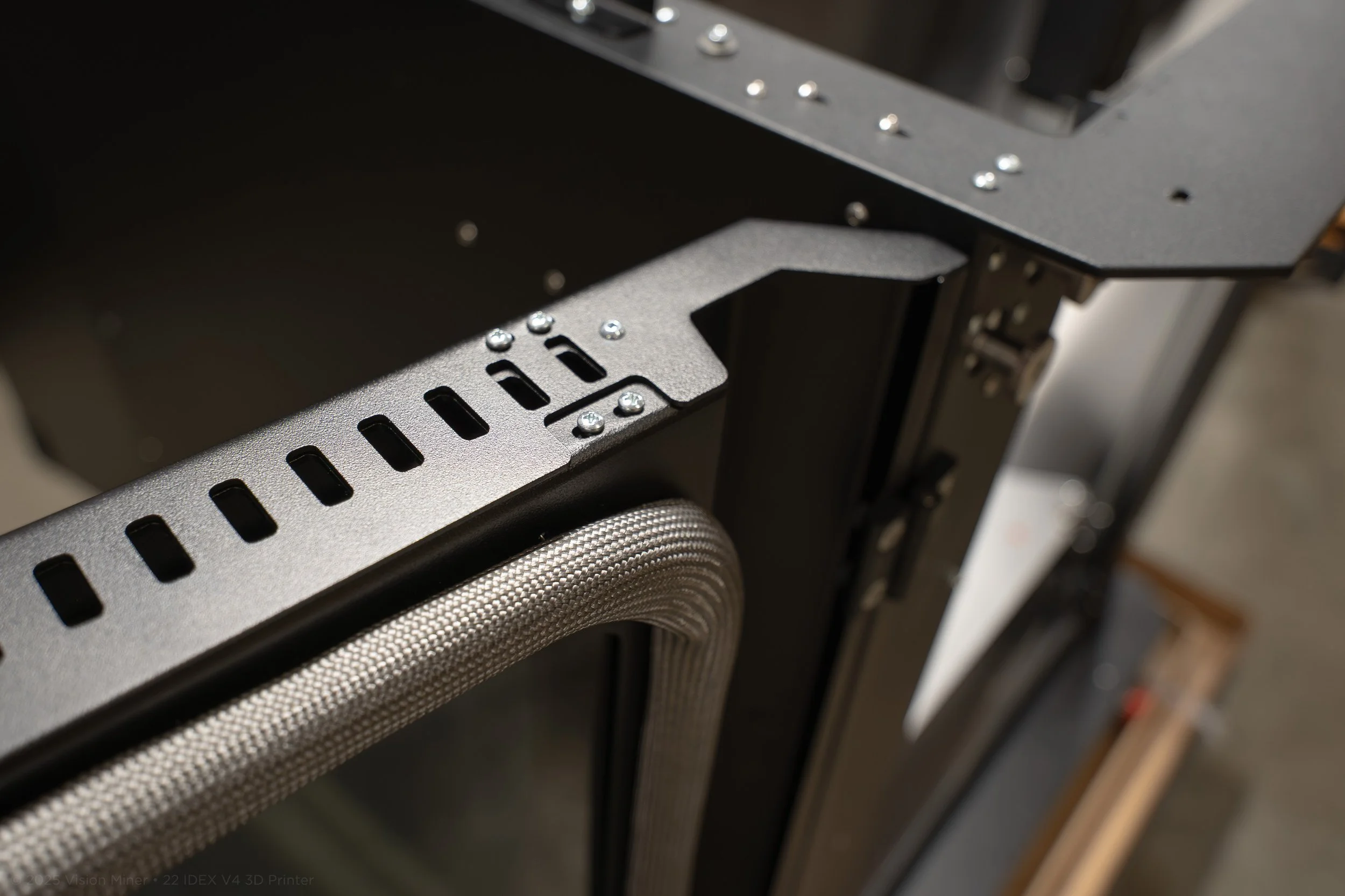 Close-up view of a metal frame with perforated slots, screws, and a braided cable attached, part of a 3D printer or electronic equipment.