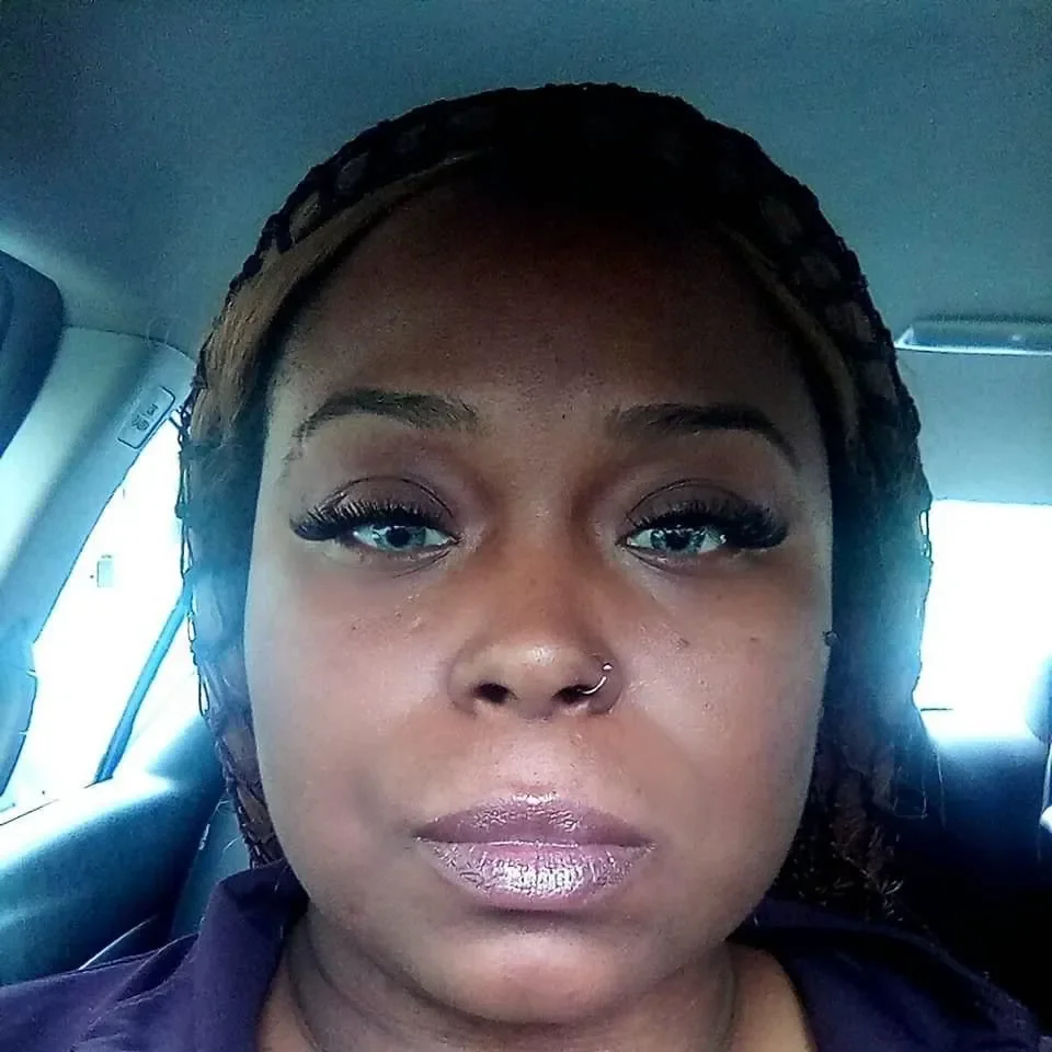 Close-up portrait of a woman with a neutral expression, wearing makeup with long eyelashes and a nose piercing, inside a vehicle.