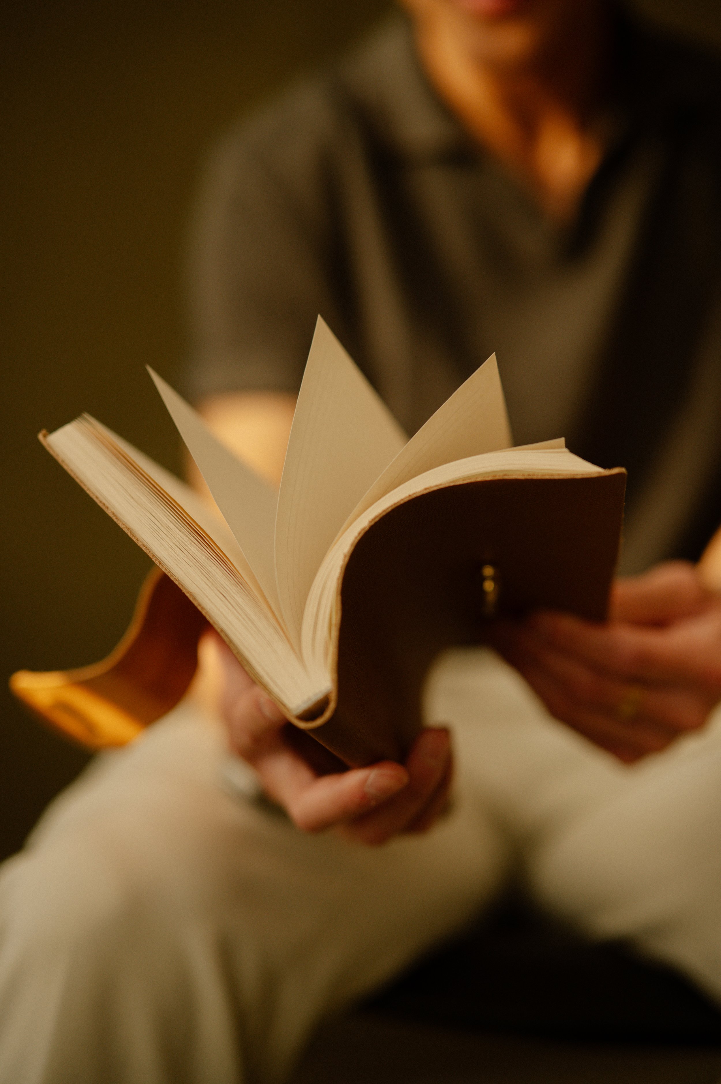 Person reading a hardcover book with a dark cover and cream-colored pages, sitting in a softly lit environment.