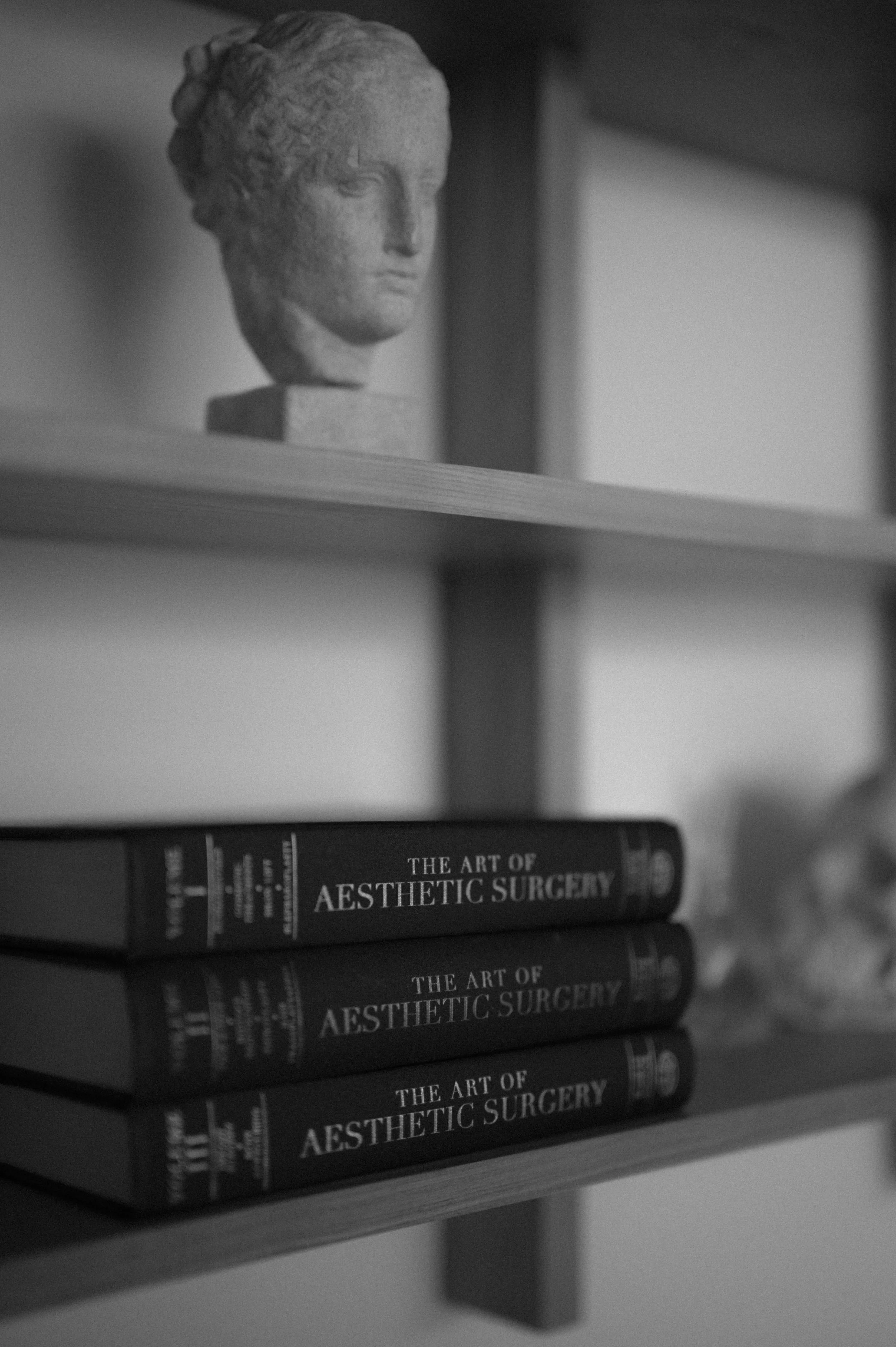 Black and white photo of a shelf with three copies of a book titled 'The Art of Aesthetic Surgery' stacked horizontally and a bust sculpture of a woman's head on the upper shelf.