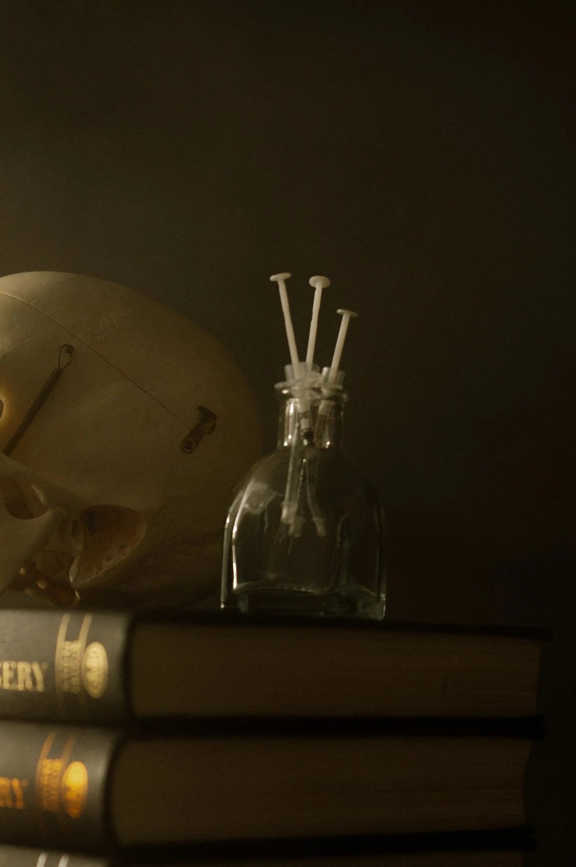 A skull with a large syringe in its mouth, a glass bottle with three syringes on top, and two closed books with dark covers.